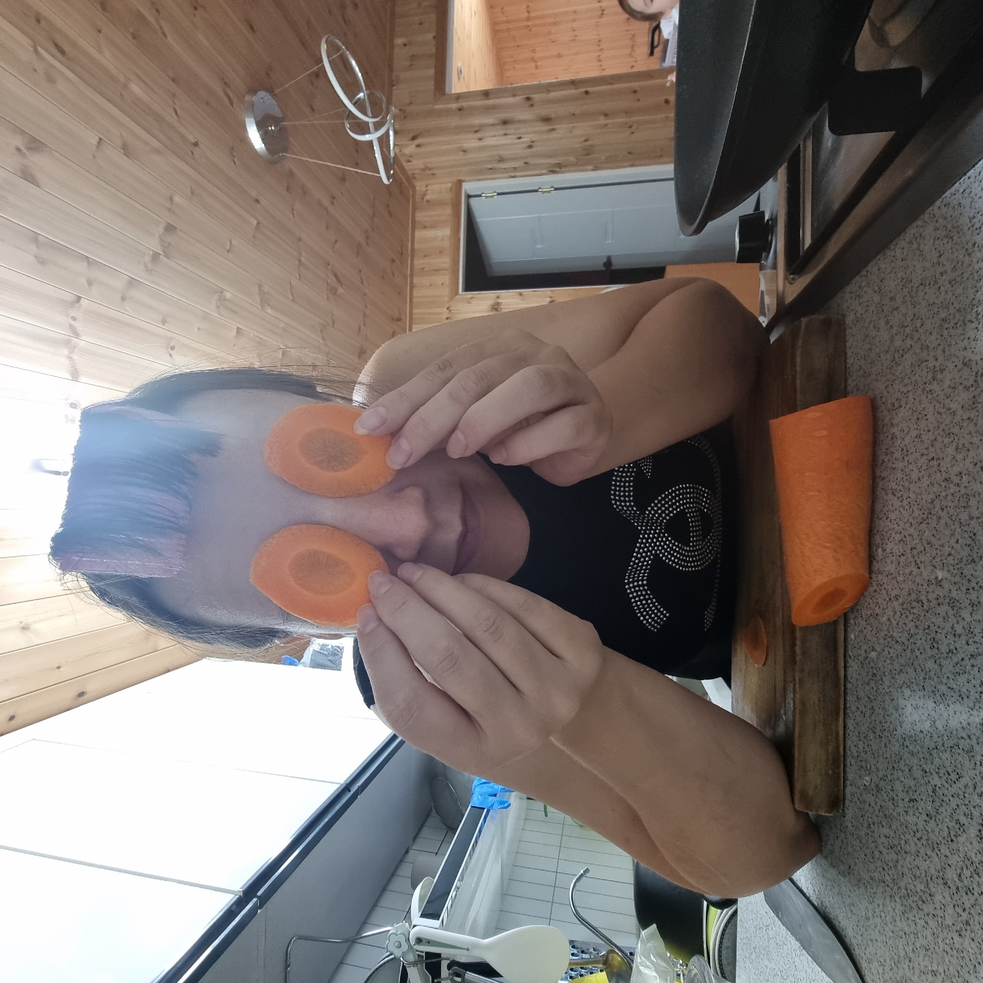 A woman with pink hair rollers is holding two slices of carrot over her eyes in a kitchen. She is wearing a black t-shirt with a sparkly design. In front of her is a cutting board with a partially cut carrot and a knife. To her right, there is a large black frying pan. The kitchen has light-colored countertops and wooden paneled walls and ceiling. The scene is indoors, likely in a residential setting in Dunnae-myeon, South Korea.