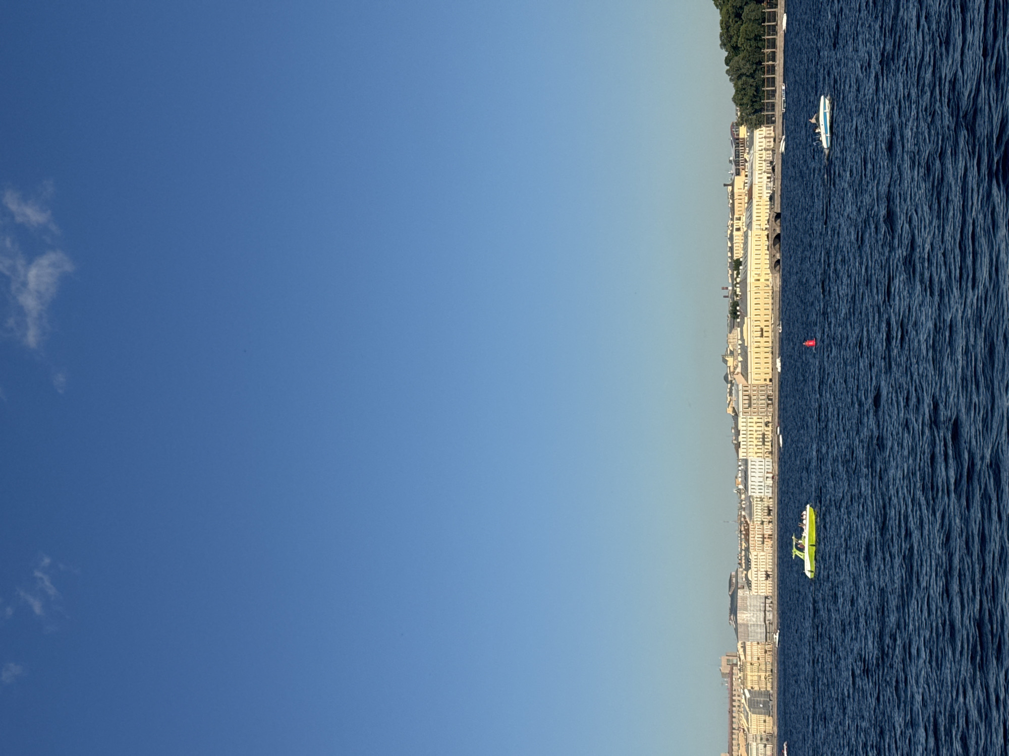 The image depicts a wide, deep blue body of water, likely a river or canal, under a clear, pale blue sky with a few wispy clouds. The water is moderately choppy, with visible ripples and waves. In the foreground, the water dominates the scene.
Further in the distance, along the horizon, a long, elegant row of light-colored buildings with ornate architecture lines the waterfront. These buildings appear to be historic, with many windows and varied rooflines. Some appear to be pale yellow, while others are white or cream. A line of trees with dark green foliage forms a backdrop to the buildings on the far right of the frame.
Several boats are present on the water. Most notably, a bright lime green and white speedboat is positioned in the mid-ground, carrying several people. To the right of this boat, a smaller white motorboat with blue trim is also visible, with one person on board. Further in the distance, other smaller boats are scattered across the water, and a red buoy is visible. The overall scene suggests a sunny day in a European city with significant waterways and historical architecture, consistent with Saint Petersburg, Russia, specifically along the Neva River.