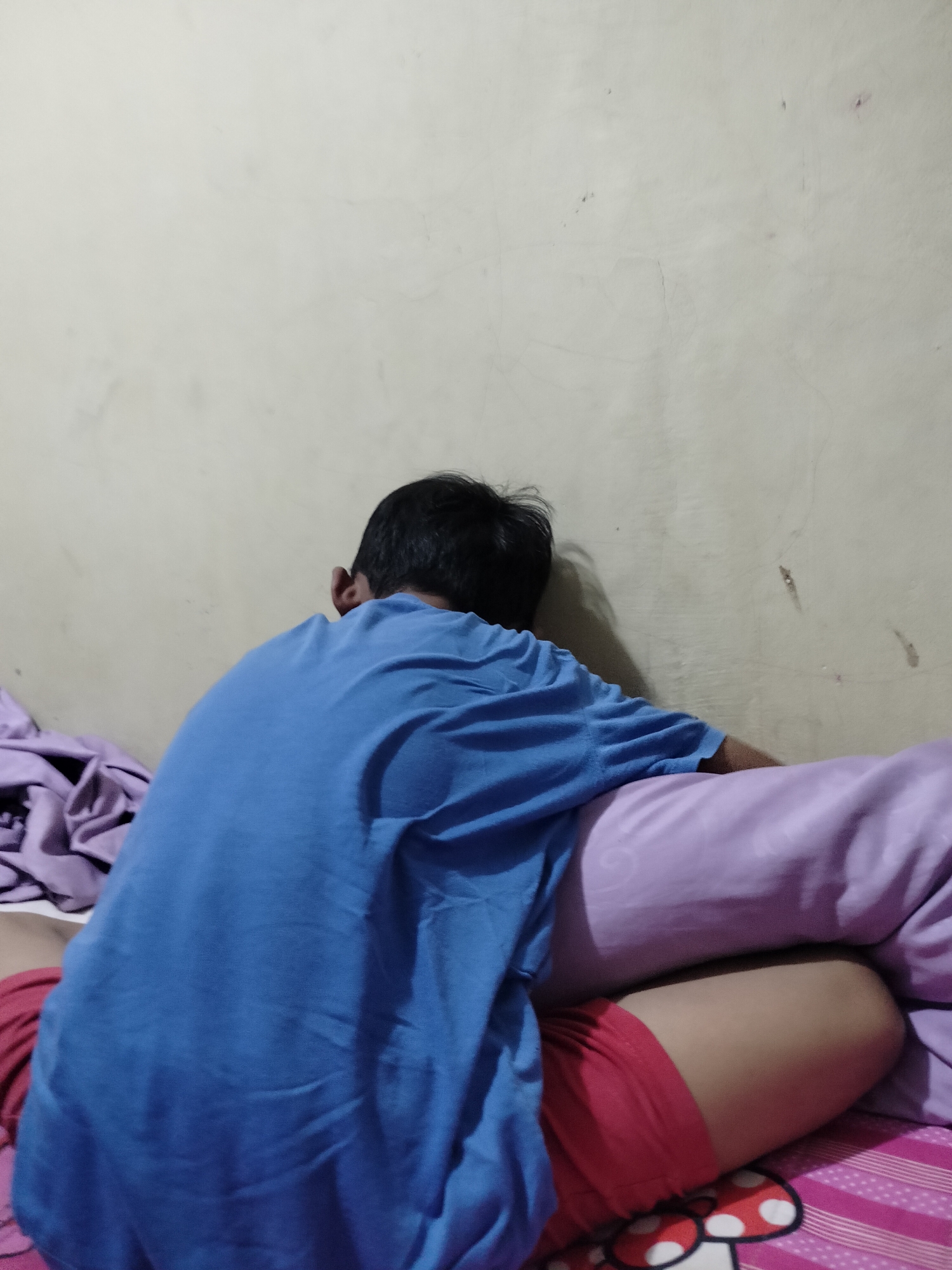 The image depicts a person lying on a bed in what appears to be a bedroom. The person is wearing a blue t-shirt and red shorts. They are positioned with their back to the camera, facing a wall. Several pillows and blankets are visible on the bed, some of which are purple and pink. The wall behind the person is a light, neutral color and shows some signs of wear, including faint stains and hairline cracks. The bed appears to be covered with patterned sheets. The overall impression is of a domestic indoor setting. The location within Kumbangindah, Indonesia, is not discernible from the visual information provided.