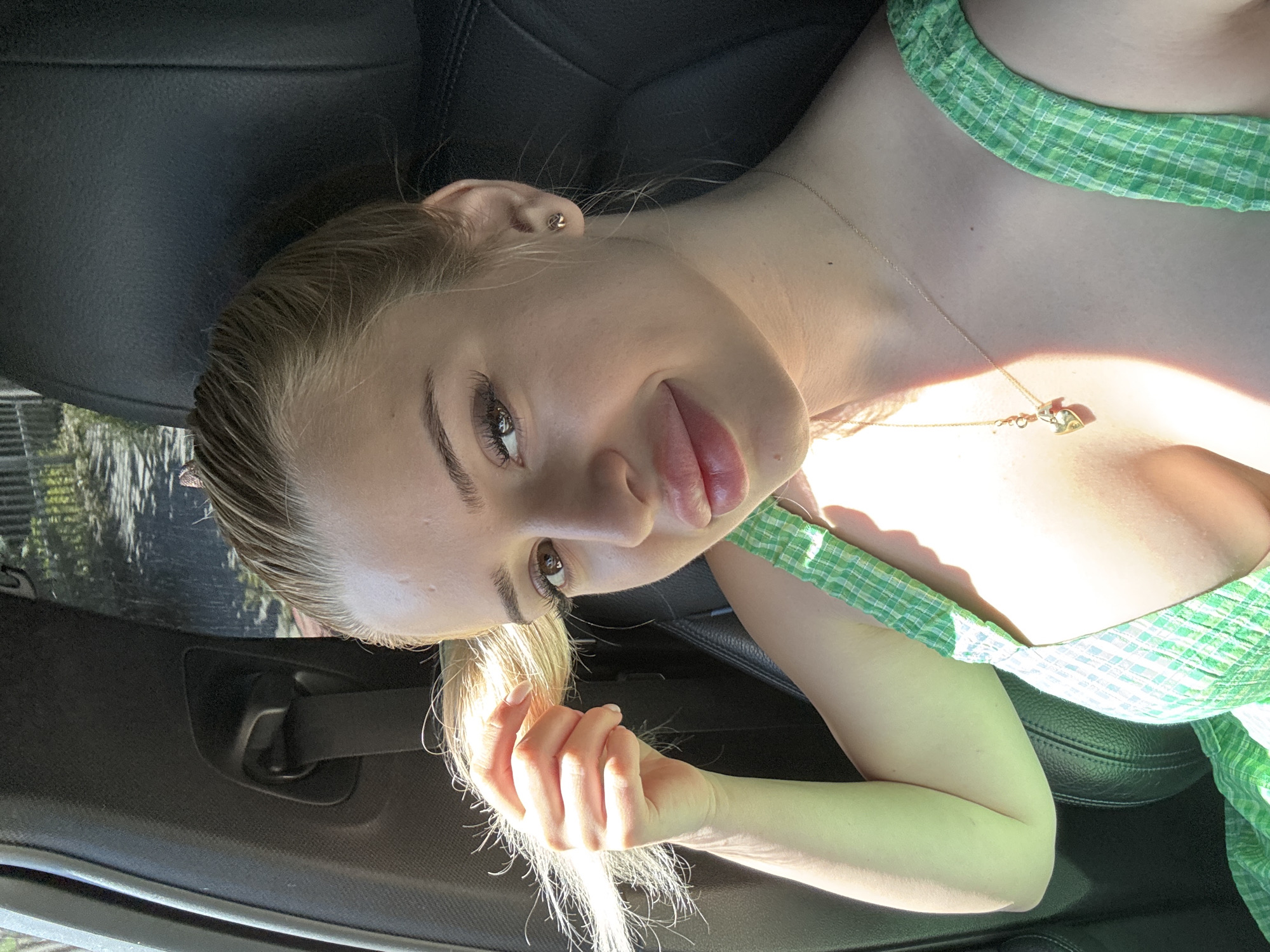 A young woman is pictured inside a car. The camera angle is tilted. The woman has light skin, blonde hair pulled back, and is wearing a green and white plaid dress with a gold necklace. Her left hand is visible in the foreground. The background consists of the car's interior, including the black leather seat and interior details. The environment suggests daylight, with sunlight casting shadows across the scene. The location is indeterminable from the image, so the city of Byala Slatina, Bulgaria cannot be confirmed.