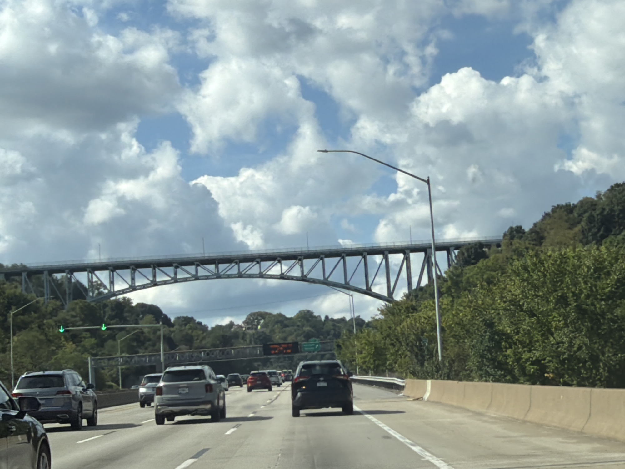 The image shows a view from a vehicle driving on a multi-lane highway in Pittsburgh, Pennsylvania, United States of America. The highway has concrete barriers along the side and traffic lights are visible in the distance. A large steel truss bridge spans across the highway, with trees and foliage covering the surrounding hills. Several vehicles, including SUVs and cars, are traveling on the highway. The sky is partly cloudy with blue patches visible between the clouds. The overall scene depicts a typical highway commute under a cloudy sky with a prominent bridge in the background.