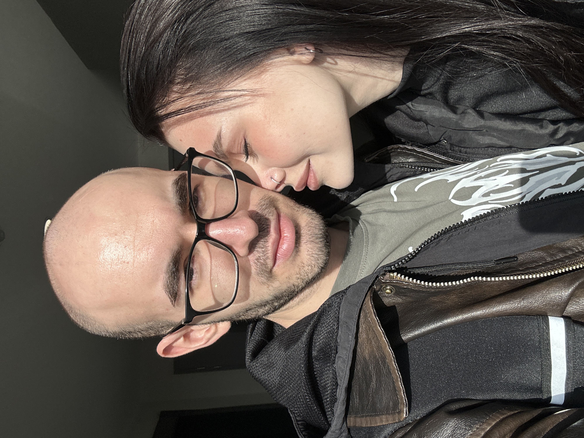 The image depicts a close-up of a man and a woman. The man is bald with a light complexion and stubble. He is wearing black-framed glasses and appears to be wearing a green t-shirt with a white graphic design. He is also wearing a dark brown leather jacket with a black hooded jacket layered beneath. The woman has long, dark hair and fair skin. She is resting her head against the man's head, with her eyes closed. She is wearing a dark jacket. The photo is taken indoors with natural light illuminating the subjects. The location cannot be determined from the image.