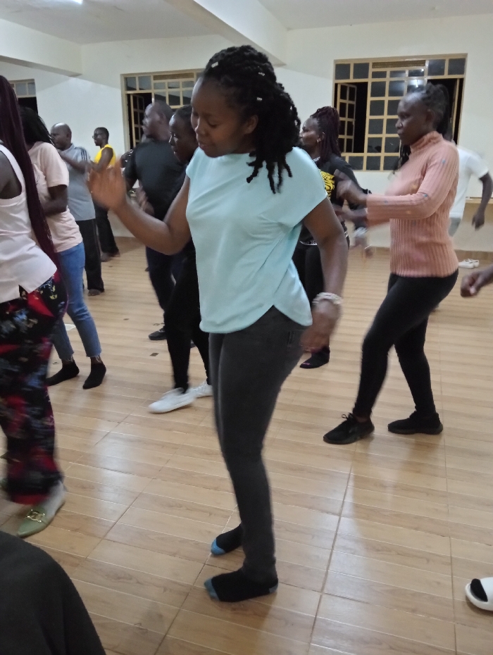 The image depicts an energetic indoor scene at Riara Ridge, Kenya, featuring a group of people engaged in what appears to be a dance or group exercise session. The central subject is a woman with dark dreadlocks, captured in mid-movement. She wears a light blue short-sleeved top and dark gray skinny jeans, with black socks, one of which has a light blue toe. Her arms are slightly raised, and her body is oriented towards the right, indicating active participation in the activity.
To her right, another woman with dark hair, wearing a peach or light orange long-sleeved ribbed top and black leggings, is also seen moving with a similar posture. On the far left, partially cropped, is a person seen from behind, wearing a light-colored top and patterned pants. Several other individuals, men and women, are visible in the background and foreground, many blurred due to motion or shallow depth of field, suggesting a larger gathering. One man in the background is wearing a yellow shirt.
The setting is a spacious room with light cream-colored walls and a light brown tiled floor. Windows with dark grilles are visible in the background, suggesting natural light might typically enter, but the primary illumination in the image appears to be artificial. The overall mood is active and communal. No visible text is present in the image.