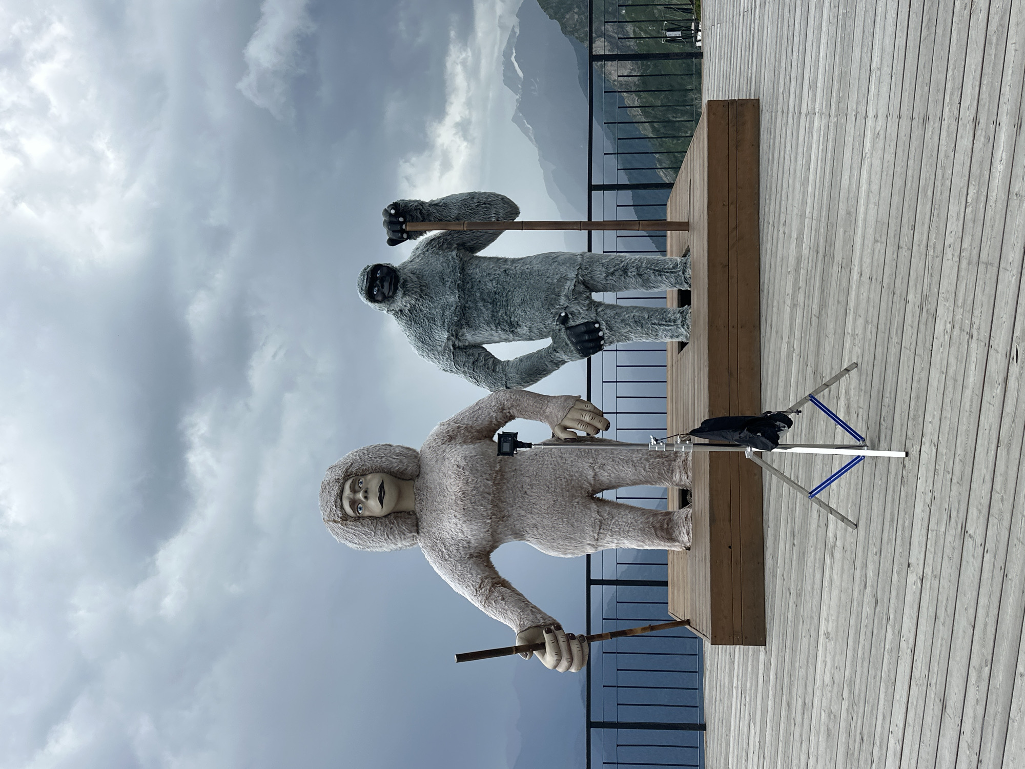 Two life-sized, anthropomorphic statues, resembling yetis or Bigfoot creatures, stand on a wooden platform overlooking a mountainous landscape under a cloudy sky. The statue on the left is lighter in color with a fuller, curly headdress and is holding a wooden stick. The statue on the right is grey and shaggier, also holding a wooden stick. In the foreground, a tripod with a black bag is set up on a weathered wooden deck. A metal railing separates the deck from the steep drop-off of the mountain. The scene is situated in Estosadok, Russia, likely a mountain resort area.