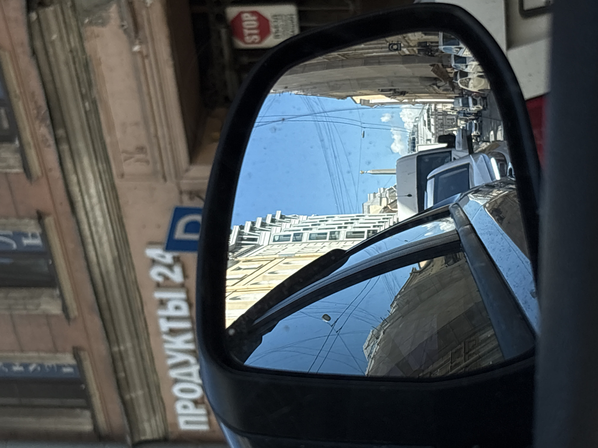 The image is a photograph taken from inside a car, looking into the side mirror. The mirror reflects a street scene in Saint Petersburg, Russia. The building in the background has signage that reads "ПРОДУКТЫ 24," indicating a 24-hour grocery store. To the right of the building, a red and white octagonal "STOP" sign is visible. The mirror also reflects several cars on the street, suggesting traffic. The sky is blue with some clouds. The overall impression is of a typical urban street in Russia.