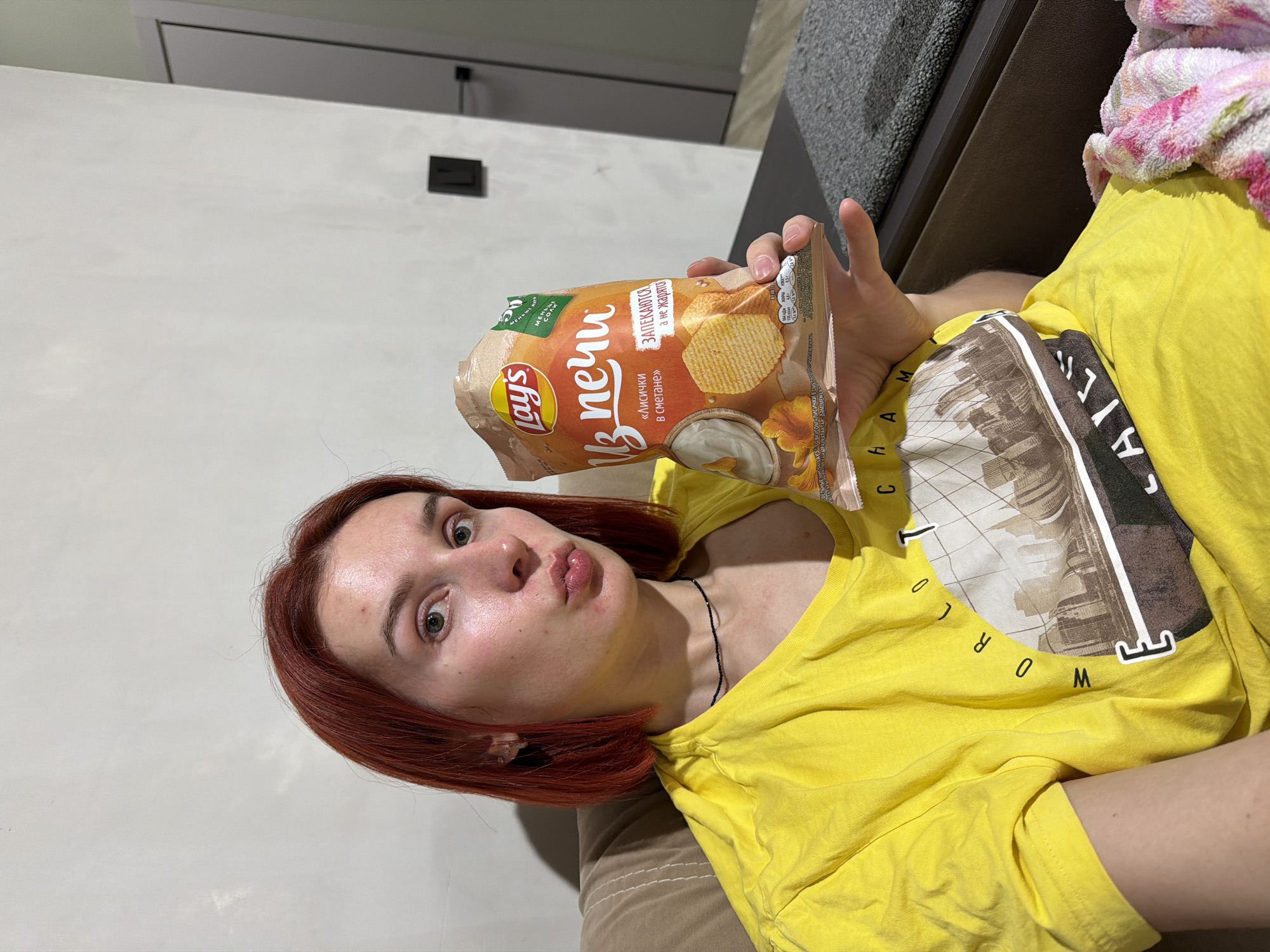 A woman with red hair is holding a bag of Lay's chips in front of her face. She is wearing a yellow t-shirt. She is lying on a brown couch. The bag of chips is Lay's "From the Oven" with a sour cream and chanterelle mushroom flavor, judging by the Russian text on the bag. A portion of the text on the t-shirt is “WORLD”. The image appears to be taken indoors, possibly in a living room. The bag of chips indicates the location might be Russia. The absence of further contextual information within the image prevents identification of the precise location within the city of Chaikovsky.