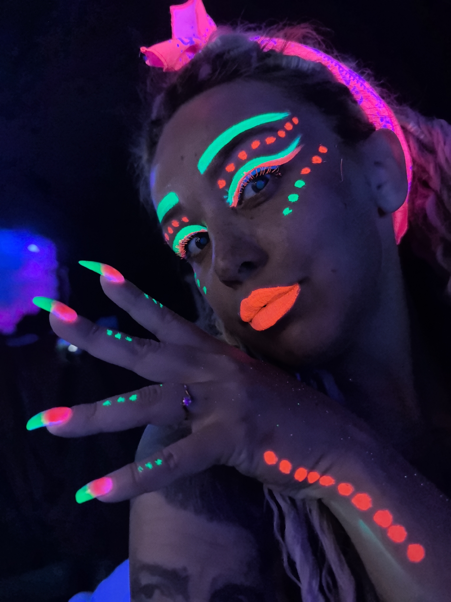 A woman with neon makeup and nail polish is photographed in a dark environment under UV light. Her makeup includes green stripes above her eyebrows, green and orange details around her eyes, and bright orange lipstick. She also has green dots on her fingers, and a line of orange dots on her arm. Her nails are long, with a green tip and pink base. She is wearing a pink headband with a bow. She is wearing a ring on her finger. The location of the city of Noviy, Russia is not evident from the image.