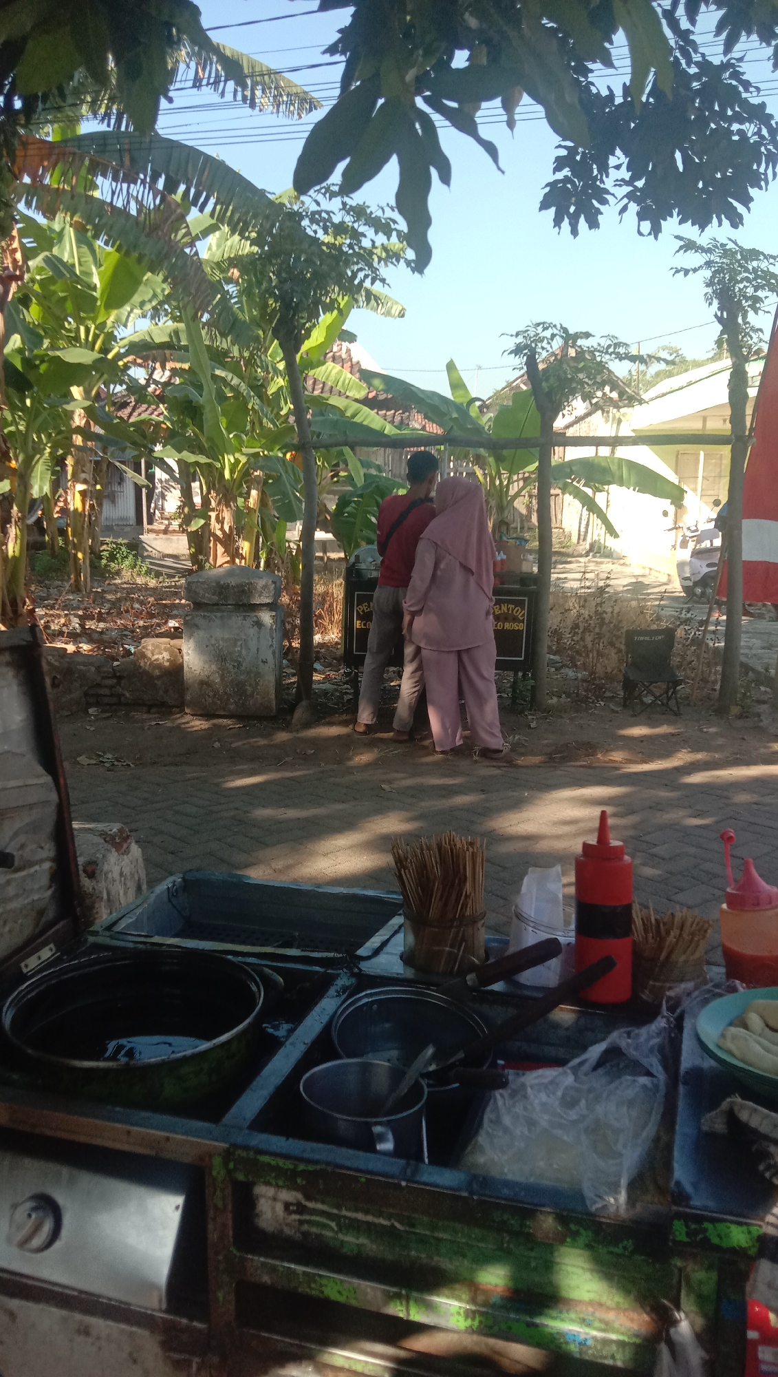 The image depicts an outdoor scene in Ngempit, Indonesia, likely a street vendor's stall. In the foreground, a food cart is visible, containing various cooking implements such as pots, pans, and a container of skewers. There are also condiment bottles, including a red squeeze bottle and a pink squeeze bottle. In the midground, two individuals, a man wearing a red shirt and grey pants, and a woman wearing a pink hijab and matching outfit, are standing by a wooden structure. Behind them, a sign is partially visible with text that appears to be in Indonesian, possibly advertising a food product. The background reveals a tropical environment with lush banana trees and some houses. The ground is paved with interlocking bricks. The lighting suggests it is daytime, with clear sky visible.