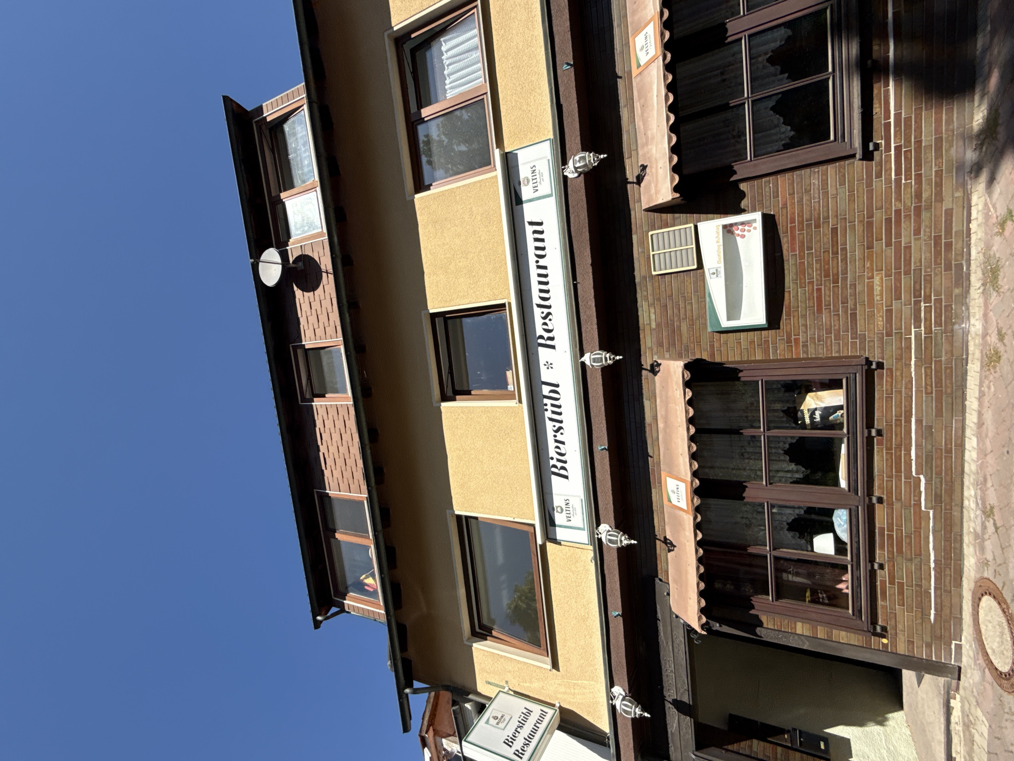 The image shows the exterior of a restaurant in Bad Sachsa, Germany, on a clear, sunny day. The building is made of brick on the lower level and a light yellow stucco on the upper levels. A sign above the entrance reads "Bierstübl * Restaurant" with Veltins branding visible. There are awnings over the windows on the lower level, and several windows are visible on different floors. The sky is a bright, clear blue.