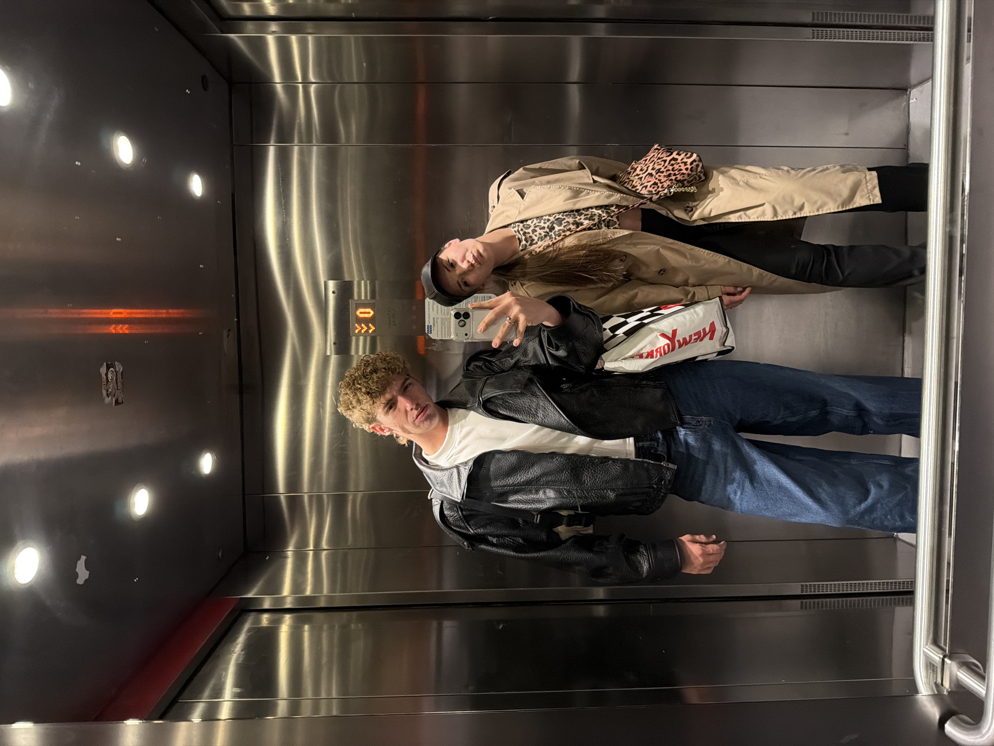The original image is a selfie taken inside a modern elevator with brushed metal walls, rotated 90 degrees clockwise. Two individuals are depicted, seemingly posing for the camera.
The person in the foreground, positioned on the right side of the frame (if viewed upright), is a young man with short, curly, light brown hair. He wears a white t-shirt, a black leather jacket, and blue jeans, looking directly at the camera with a neutral expression. He holds a smartphone, likely an iPhone, pointed towards himself and the other person, indicating he is taking the photo.
Behind him, on the left side of the frame (if viewed upright), is a young woman with long, dark hair. She is dressed in a beige trench coat, a leopard print top, black pants, and a black beret. A leopard print handbag is slung over her shoulder. She also looks towards the camera, making a peace sign with her fingers.
The elevator interior features bright, circular recessed lights on the ceiling, casting reflections on the metallic surfaces. An orange horizontal light strip is visible on the left wall. On the right wall, a control panel displays "0" with an upward arrow and three small dots, possibly indicating the current floor and movement direction. Between the two individuals, a white and black checkered shopping bag with red text is partially visible. The text on the bag includes "NEW YORK" and "ЯРОШ" (Yarosh), likely referencing a brand.
The activity is a casual selfie, capturing the two individuals together in an elevator, possibly while traveling or waiting. The artificial lighting suggests an indoor setting, and no specific time of day is discernible from the ambient light. The context provided indicates the location is Sofia, Bulgaria.