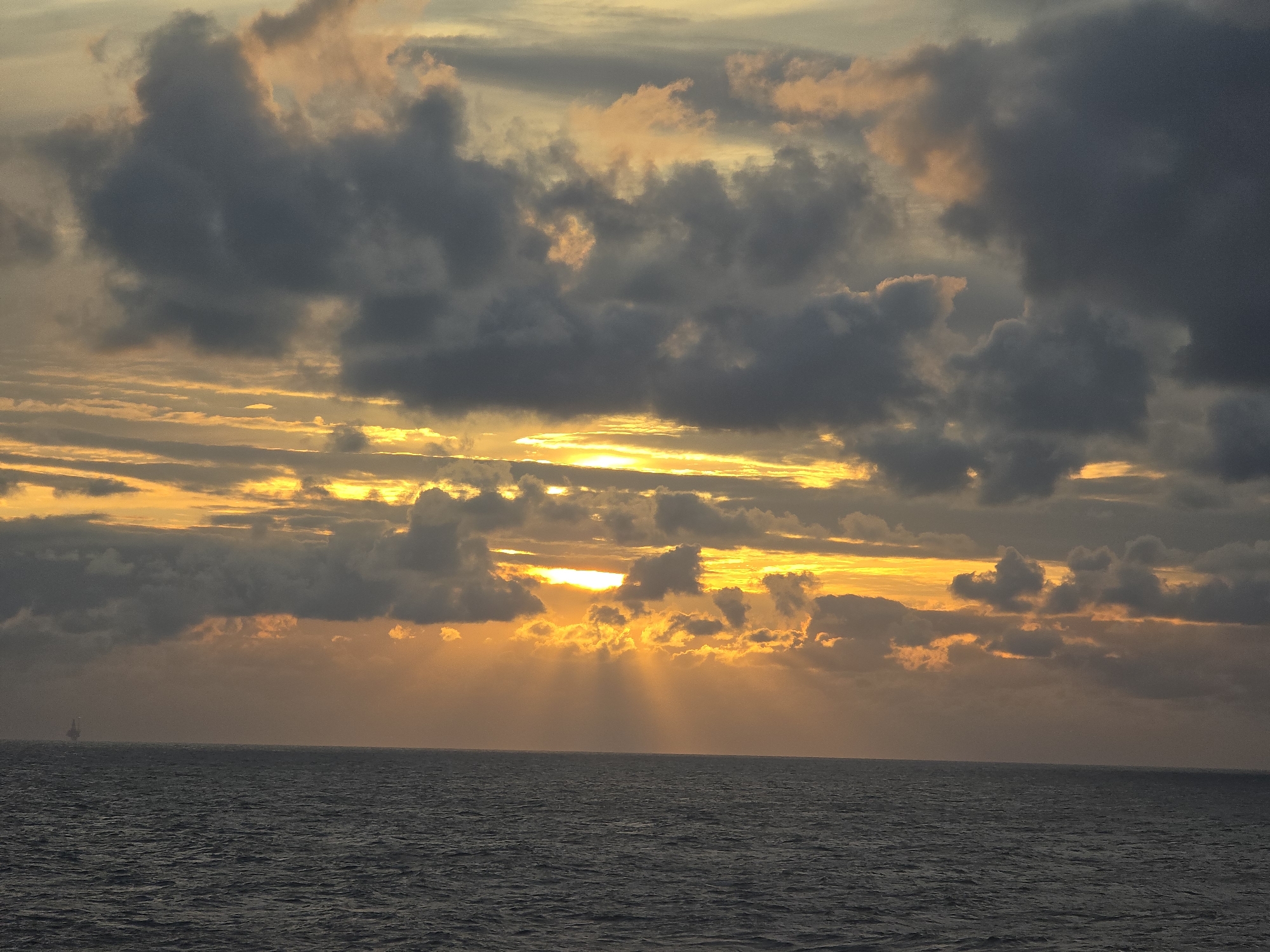 A wide-angle shot captures a sunset over the ocean. The sky is filled with clouds of varying shades of gray, illuminated by golden sunlight piercing through openings. A set of sunbeams can be seen going from the clouds to the water. On the horizon, to the left, a distant structure sits upon the water, appearing to be an oil rig. The sea itself is a dark, flat expanse reflecting the dimming light. The location is Unknown, Unknown.