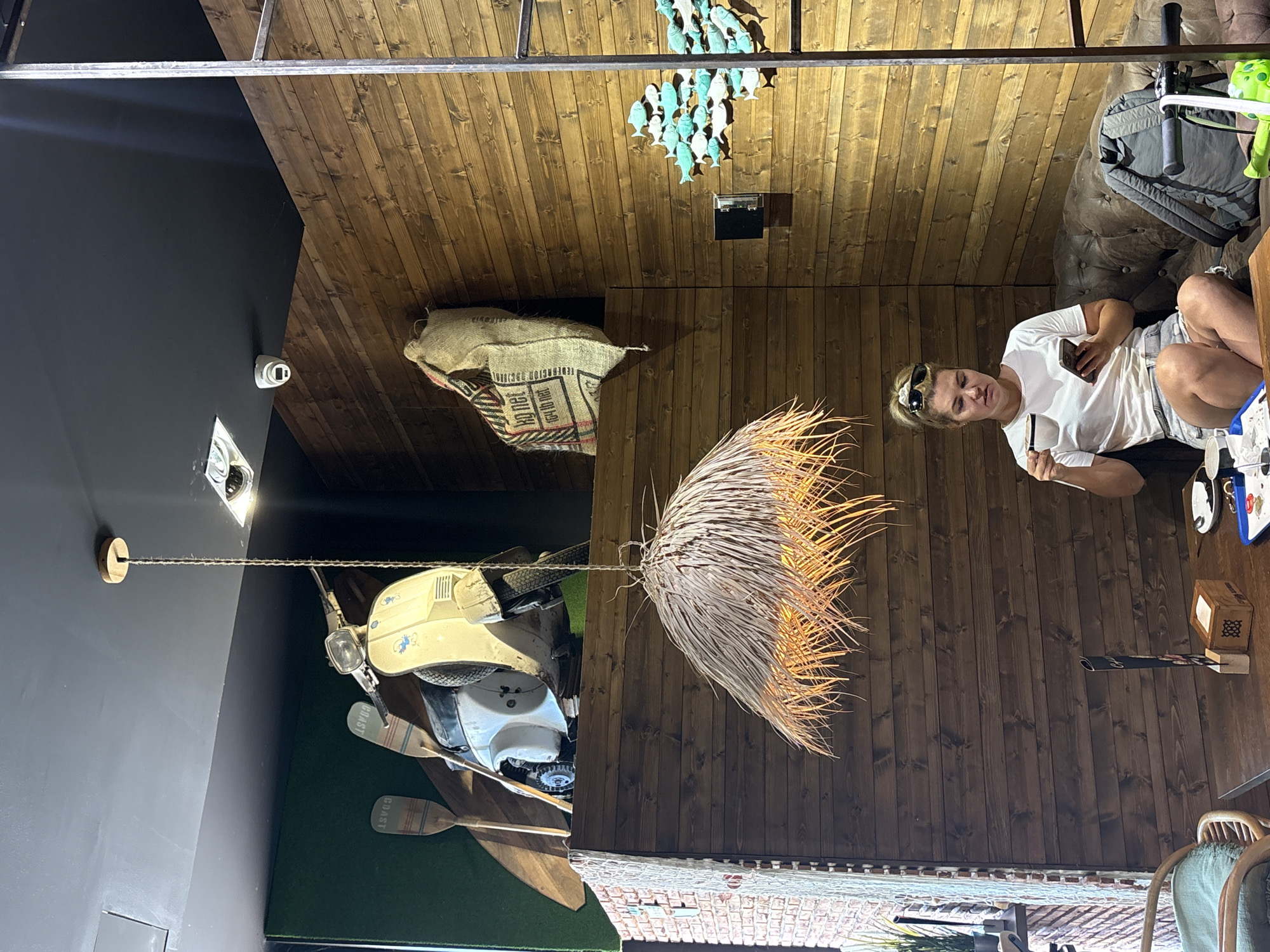 The image depicts an indoor scene with a rustic, wooden-paneled ceiling and walls. A woman is seated on a brown leather couch, holding a phone and a small object in her hands. She is wearing a white t-shirt and striped shorts, with sunglasses perched on her head. Next to her is a green backpack and a light green object.
The scene features several interesting objects. Hanging from the ceiling is a decorative mobile of small, light blue fish. Near the woman, a large, dried palm frond hangs down, possibly as a light fixture or decoration. In the background, a vintage-looking cream-colored scooter is suspended from the ceiling with ropes, positioned above two wooden oars with "COAST" written on them. A security camera is visible on a black wall in the foreground. The overall ambiance suggests a casual, perhaps eclectic, indoor space that might be a cafe, bar, or unique retail establishment. The lighting appears to be artificial, suggesting it could be any time of day. There is no visible indication of weather.