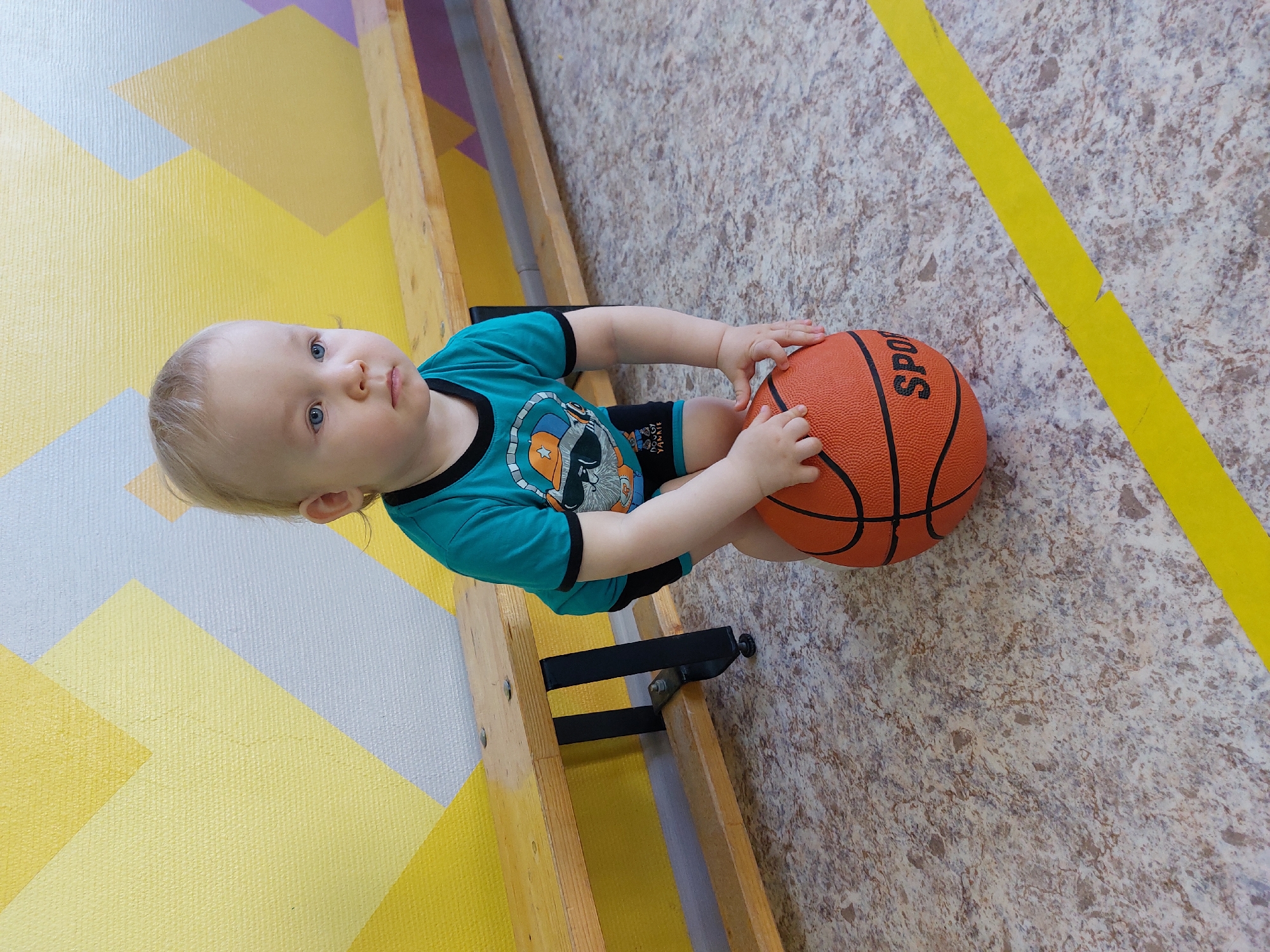 A fair-skinned toddler, wearing a turquoise t-shirt with a cartoon design and black shorts, is holding an orange basketball. The location appears to be an indoor gymnasium or play area in Chaikovsky, Russia, based on the presence of a wooden climbing frame attached to a geometric patterned wall and a marbled floor with yellow line markings. The child is positioned between the wall and the floor, with the climbing frame to their left.