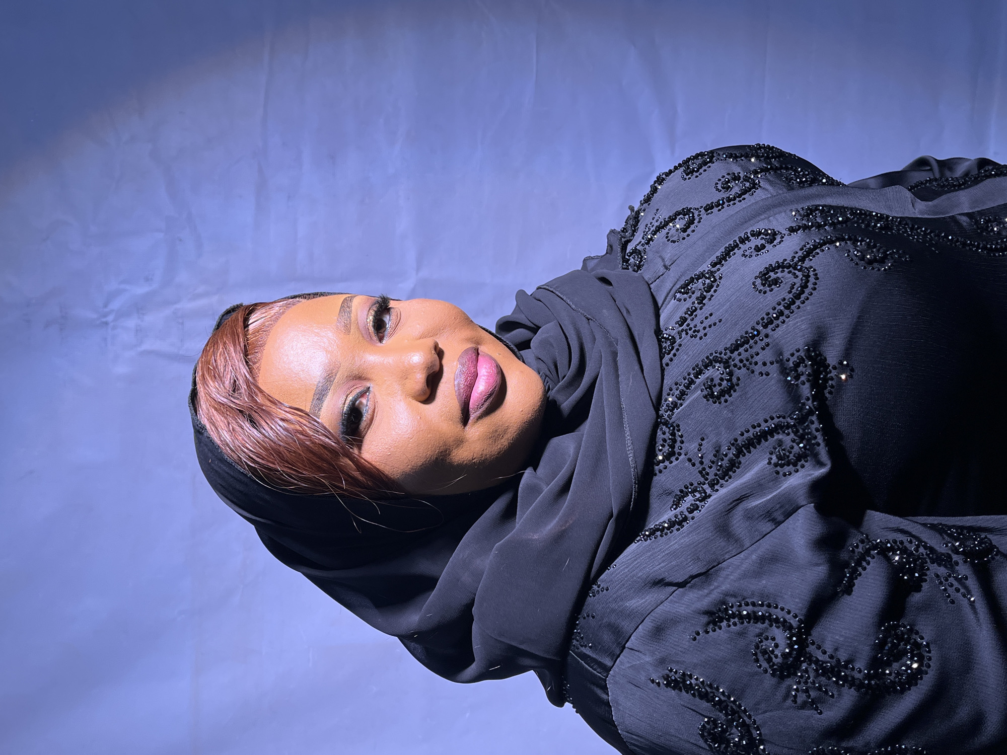 The media file shows a close-up, horizontal shot of a woman wearing a black abaya with intricate black beadwork and a black hijab. Her head is tilted, and she is looking directly at the camera with her lips slightly parted. Her makeup is visible, with dramatic eyeliner and bold lipstick. The background is a plain, textured purple backdrop. A sliver of light is visible in the top left corner, creating a spotlight effect on the background. The image appears to be taken indoors with artificial lighting. There is no visible text in the image. The scene suggests a posed portrait, possibly for fashion or personal photography, set in Barakin Akawo, Nigeria. The woman's expression is confident and alluring.