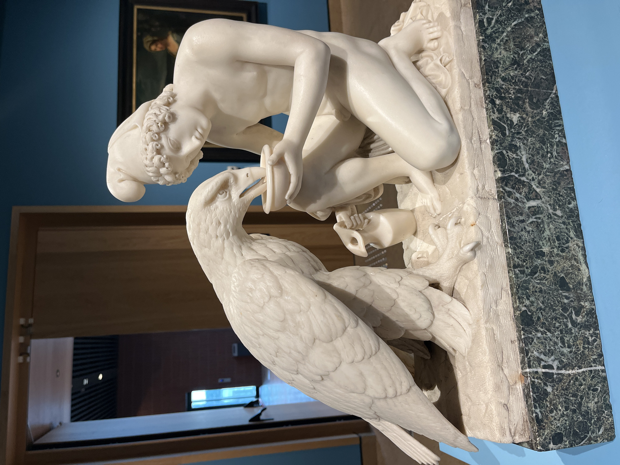 The image shows a marble sculpture depicting Ganymede feeding an eagle. Ganymede is kneeling, holding a vessel to the eagle's mouth. The sculpture is mounted on a base with a marbled green and white pattern. The sculpture is displayed in a museum setting, with a blue wall in the background and a dark-framed painting partially visible above. The location is in an unknown city and area.