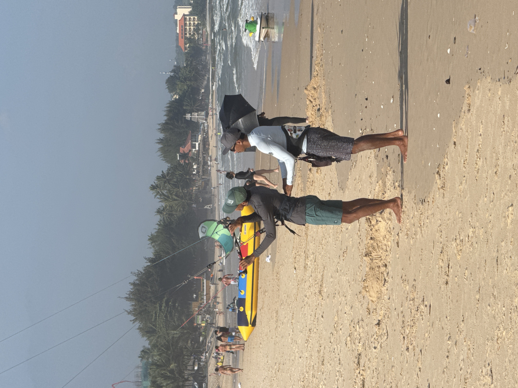 The image depicts a beach scene in Phan Thiết, Vietnam. In the foreground, two individuals are preparing for a water sport, likely kitesurfing. One person, wearing a green cap and a long-sleeved grey shirt with teal shorts, holds a green kite. The other person, to their right, wears a light blue long-sleeved shirt, a grey baseball cap, and patterned shorts, and holds a black umbrella. A yellow and blue inflatable banana boat is positioned on the sand behind the person with the kite. In the background, other beachgoers are visible, as are palm trees and buildings lining the coast. The ocean is choppy with waves. A green jet ski is also visible near the water's edge.