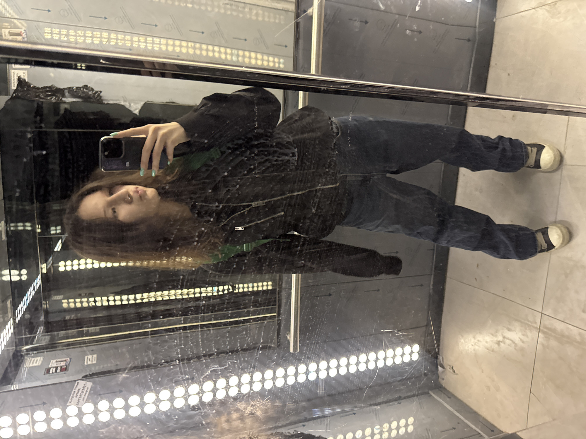 A young woman is taking a photo of herself in a mirrored elevator. She is wearing a black jacket, dark jeans, and white sneakers. The elevator is illuminated by rows of LED lights. The exact location within Unknown, Russia, cannot be determined from the image.