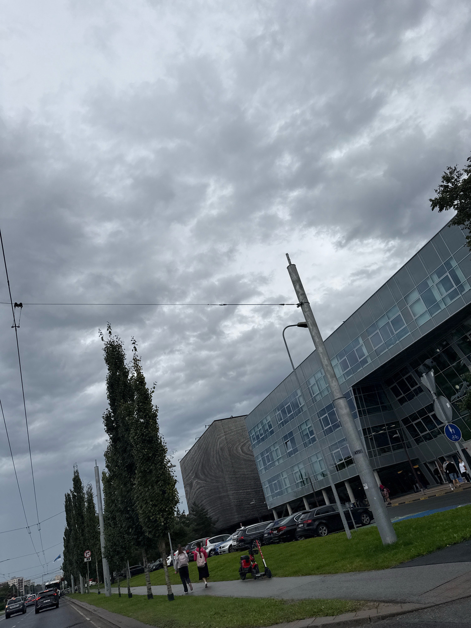 In Tallinn, Estonia, under a cloudy sky, a street scene unfolds with a modern glass-fronted building on the right, reflecting the overcast conditions. A tall, slender pole with wires attached is prominent in the foreground. To the left of the pole, a row of tall, slender trees lines a grassy area next to a street with cars. In the distance, another building with a textured, curved facade is visible. Two individuals are walking on the grass verge towards the glass building. A red electric scooter is parked on the sidewalk. The street itself features moving vehicles, suggesting an active urban environment. The overall atmosphere is one of a typical day in a European city, with architecture and infrastructure characteristic of modern urban development.