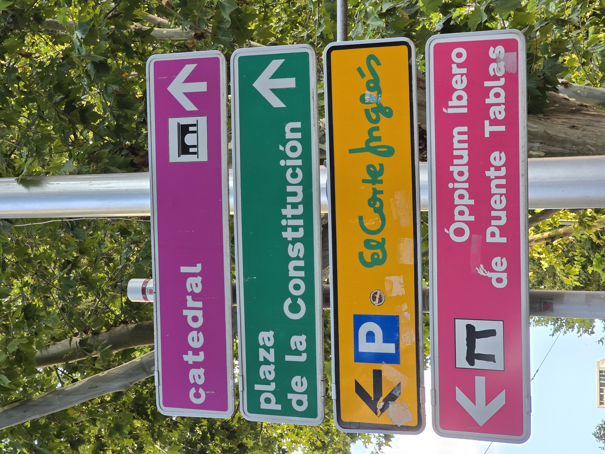 The image shows a street signpost in Jaén, Spain, with four signs indicating directions. The top sign, colored magenta, reads "catedral" with an icon of an archway and an upward-pointing arrow. Below it, a green sign reads "plaza de la Constitución" with an upward-pointing arrow. The third sign, yellow, indicates parking with a blue "P" and an arrow pointing left, and also reads "El Corte Inglés" in green script. The bottom sign, also magenta, shows an icon of a dolmen and reads "Oppidum Íbero de Puente Tablas" with an arrow pointing left. The signs are mounted on a metal pole, with trees and foliage visible in the background, and a clear blue sky overhead. There are no people or animals present in the image.