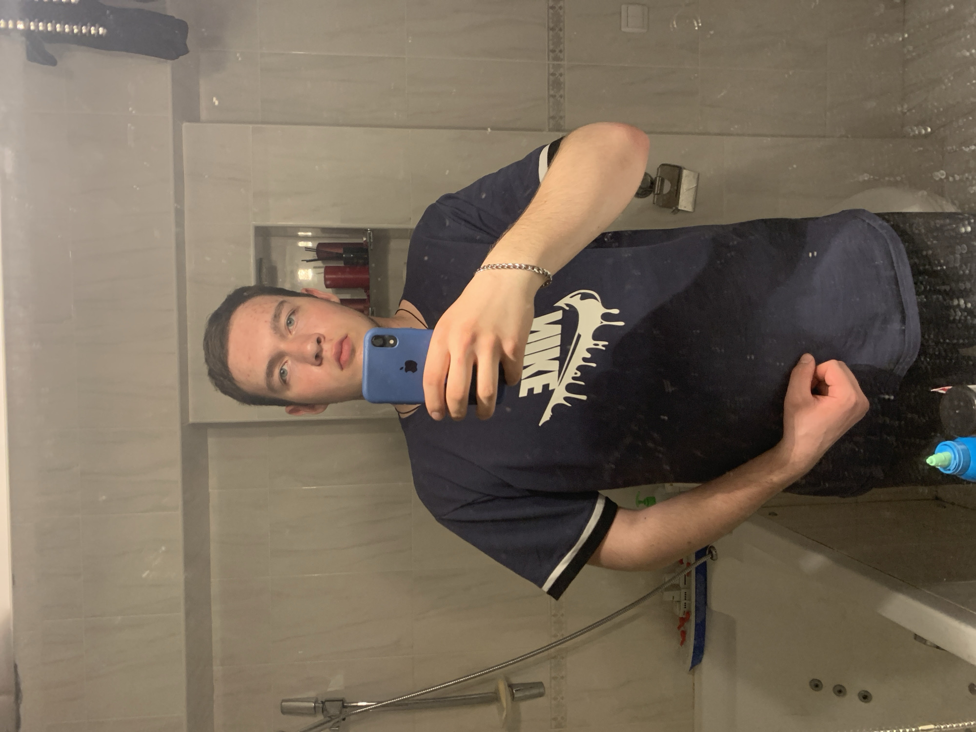 A young man is taking a selfie in a bathroom. He is wearing a dark blue T-shirt with a white Nike logo. He is holding a blue smartphone. He is wearing a silver bracelet. The bathroom has gray tile walls and a shower/tub combination. There are various toiletries visible in a recessed shelf. It is unknown if the location is in Severodvinsk, Russia.
