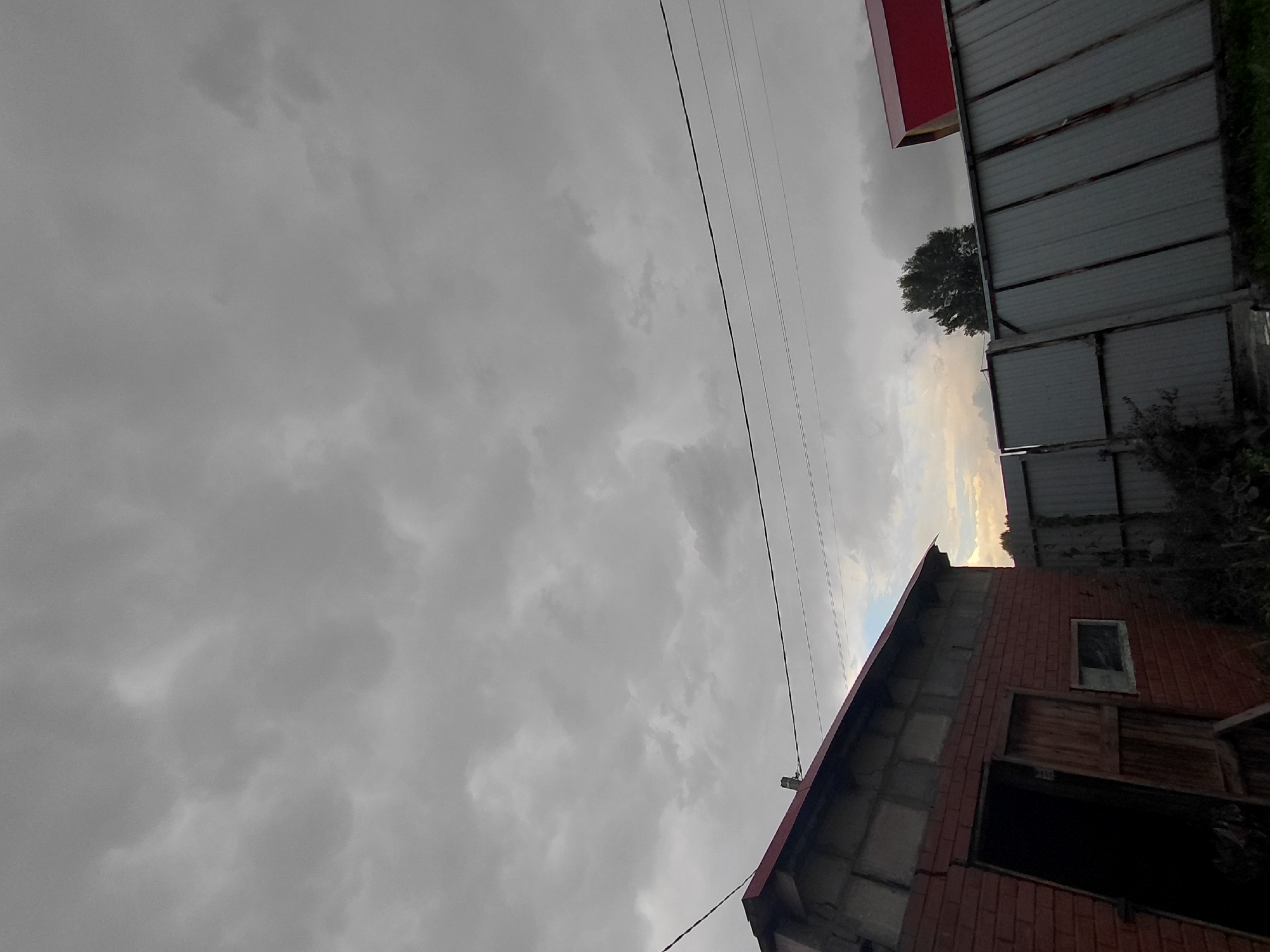 The image depicts a cloudy sky over a rural or suburban area, possibly the outskirts of Novosibirsk, Russia. In the foreground, a brick building with a red-tiled roof is visible, featuring an open doorway with wooden doors and a small window. To the right, a corrugated metal fence runs diagonally across the frame, with a glimpse of a red roof behind it. A lone tree stands in the distance. Power lines are strung across the sky. The overall atmosphere is overcast and somewhat somber.