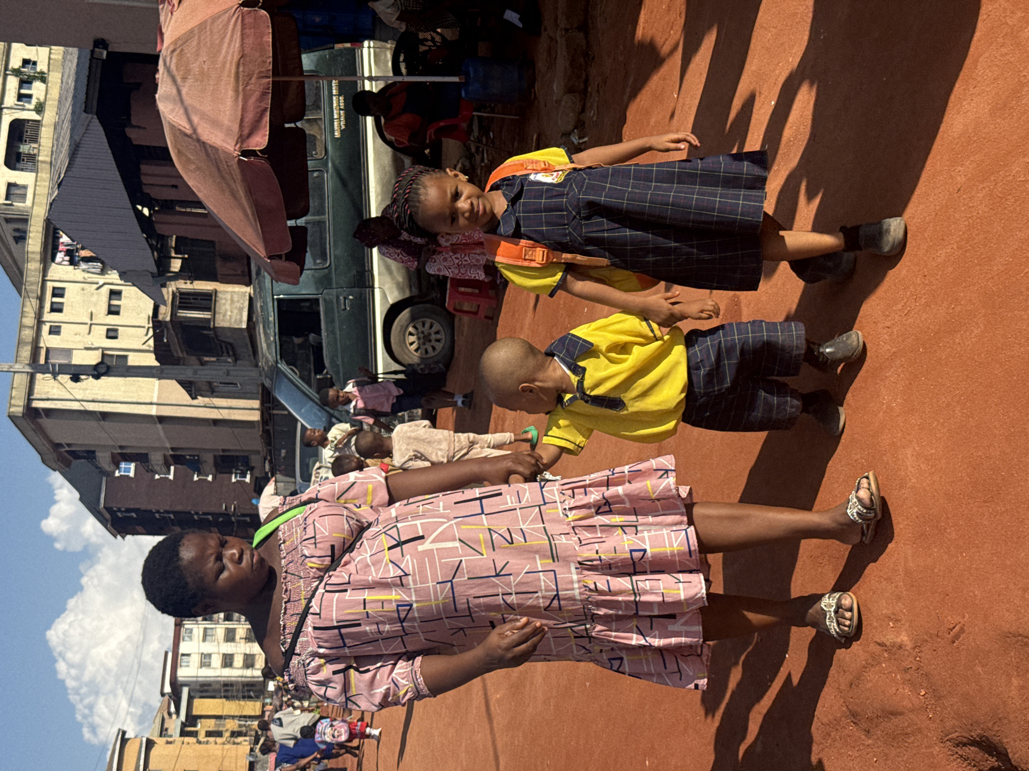 This media file captures a vibrant scene in Onitsha, Nigeria, likely during daytime. The primary focus is on a group of people, including several children in school uniforms and an adult woman.
Two young children, a girl and a boy, are dressed in blue plaid school uniforms with yellow shirts. The girl has braids and a backpack, and the boy is wearing a yellow shirt. They appear to be standing near a green van, possibly a school bus.
An adult woman is prominently featured in the foreground, wearing a pink dress with a geometric pattern. Her gaze is directed downwards, and her expression is difficult to discern due to the angle and lighting. She is wearing sandals.
The setting appears to be an outdoor, possibly bustling area. Buildings with multiple stories are visible in the background, suggesting an urban environment. There are also hints of market activity with an umbrella visible. The ground is a reddish-brown color, and strong shadows indicate bright sunlight, suggesting it's likely mid-morning or afternoon.
The children's presence in school uniforms implies an educational context, possibly waiting for or disembarking from school transportation. The overall atmosphere seems active and everyday. There is some text visible on the side of the green van, but it is illegible.