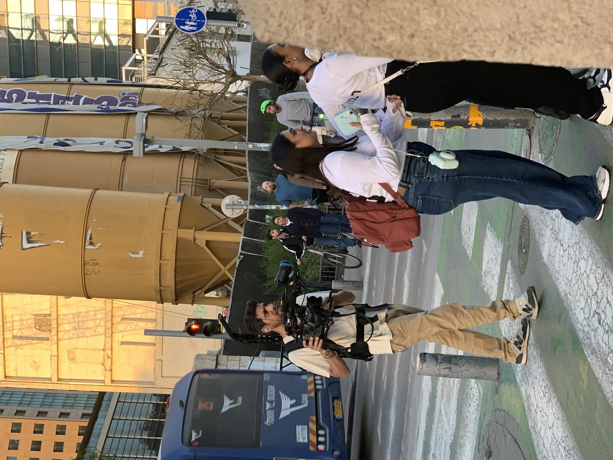 The image shows a street scene in Tel Aviv, Israel. A cameraman stands in a crosswalk, operating a professional camera rig with a blue bus behind him. Two young women, one with a blue camera on her jeans, stand facing each other in conversation. Several other people are visible in the background. The background also contains large industrial-looking tanks or silos, a building with a glass facade, and a blue bicycle and pedestrian sign. The traffic light is red.