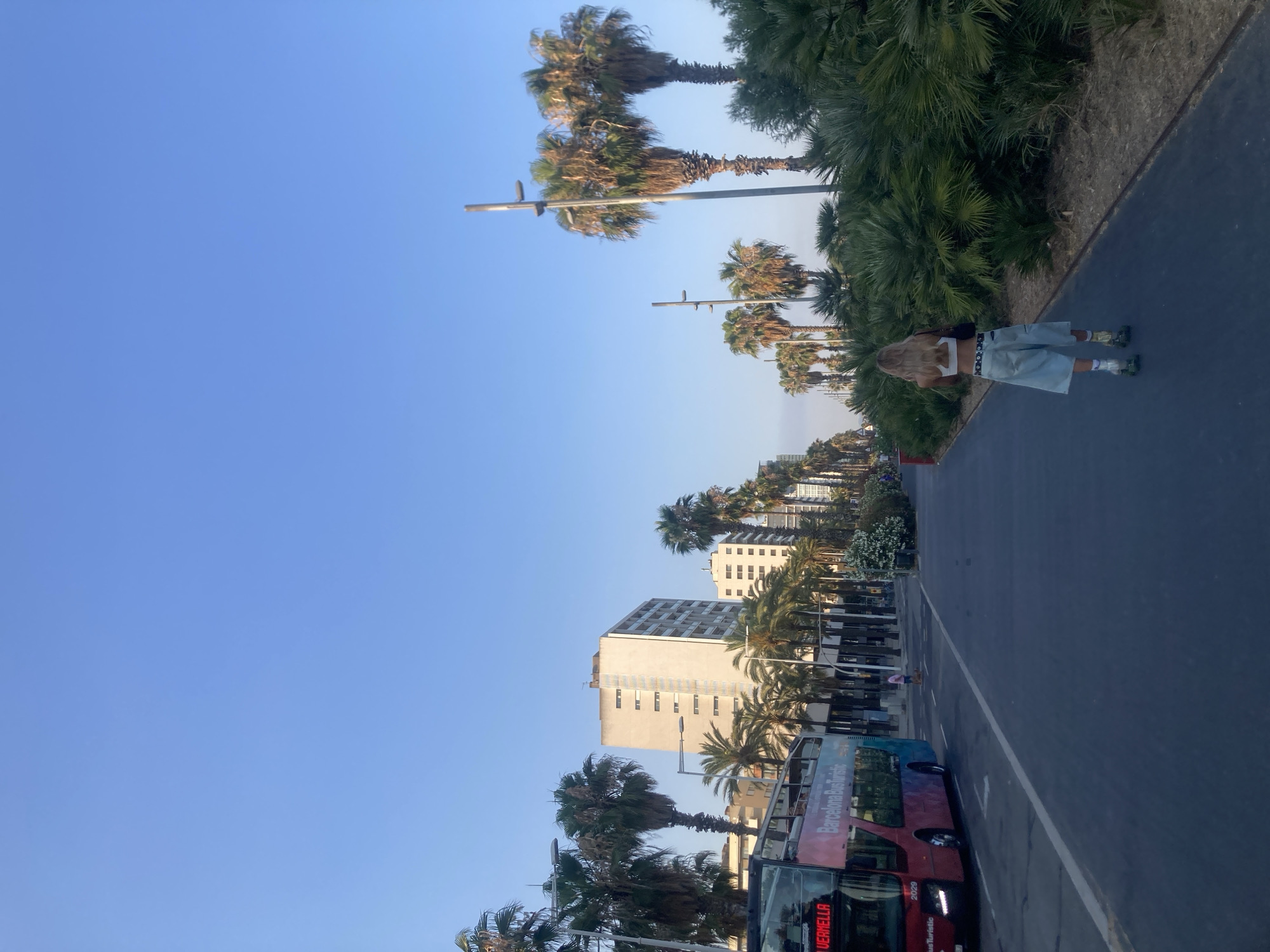 The image shows a wide, paved walkway lined with palm trees. A person with blonde hair, wearing a white crop top and baggy denim shorts, is rollerblading away from the camera on the walkway. To the left of the walkway, a red and blue double-decker bus labeled "Barcelona Bus Turístic" is driving on a street. In the background, several tall buildings and more palm trees are visible under a clear blue sky. The environment appears to be a sunny day in a coastal city, likely Barcelona, Spain, given the presence of the "Barcelona Bus Turístic".