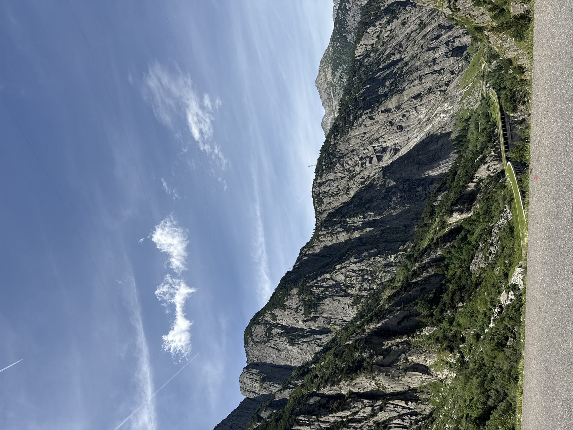 The image shows a scenic view of a mountain landscape in the vicinity of Andermatt, Switzerland. In the foreground, a paved road surface is visible. Beyond the road, a steep, rocky mountain face dominates the left side of the frame, covered in patches of green vegetation. Further into the scene, a winding road with a concrete barrier and overhead structure curves through a valley. The distant background features more mountain peaks and a bright blue sky with wispy white clouds and contrails from an airplane. The overall impression is one of a natural, mountainous environment with man-made infrastructure integrated into the landscape.