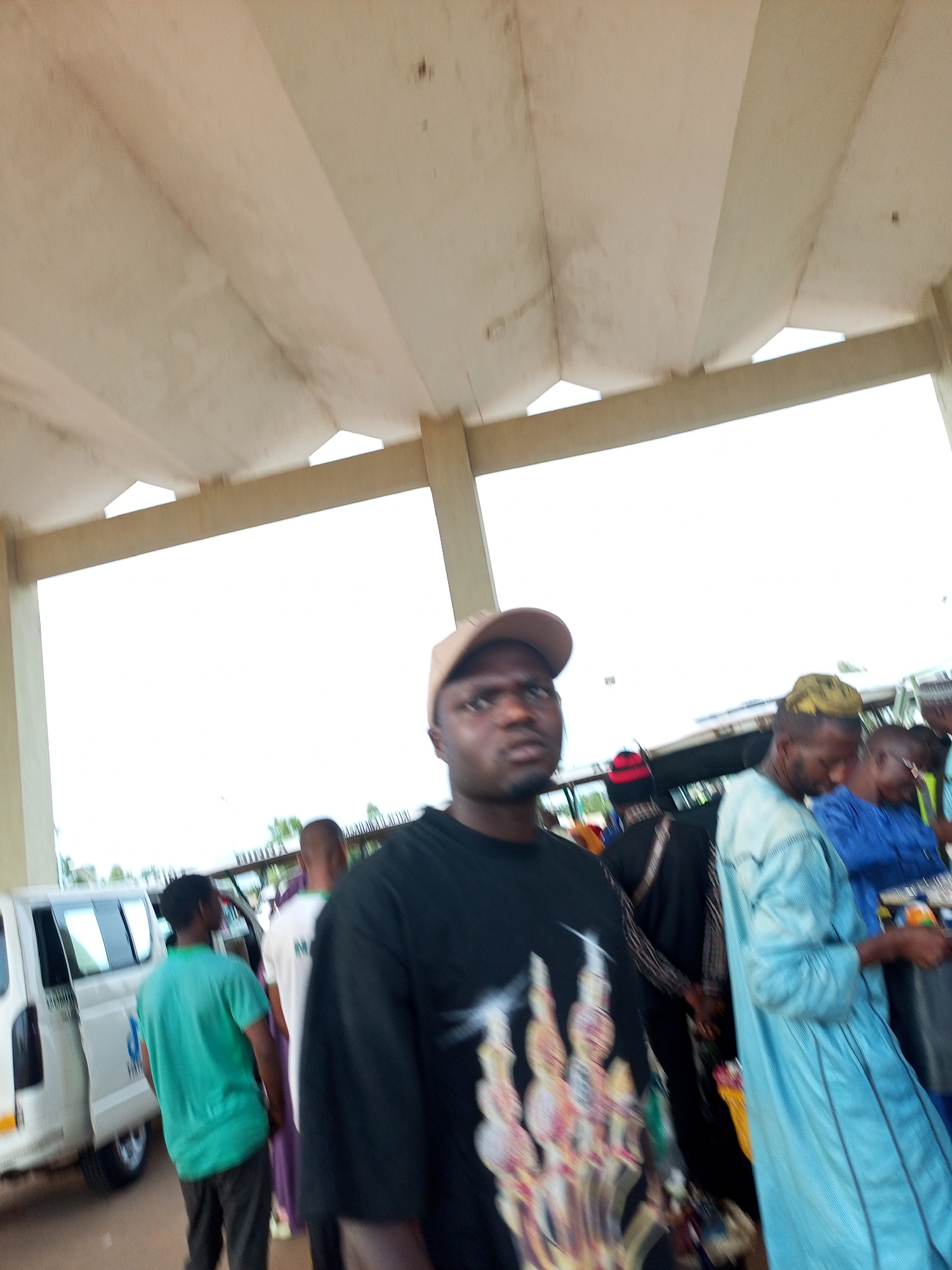 The media file shows a group of people gathered under a covered walkway, possibly a bus station or market area, in Gombe, Nigeria. The overhead structure is made of concrete beams and a light-colored ceiling, with the bright sky visible through the open sides.
In the foreground, a man wearing a black t-shirt with a graphic print and a beige baseball cap looks towards the camera with a neutral expression. To his right, another man in a light blue robe and a yellow head covering is partially visible, facing away from the camera. Further back, other individuals are seen in various clothing, including a person in a bright green t-shirt and another in a purple garment. A white van is parked on the left side of the frame.
The scene appears to be bustling with activity, suggesting a public gathering or transportation hub. The lighting indicates it is daytime, likely with clear weather given the brightness. The presence of multiple people and vehicles implies a busy environment. No discernible text is visible in the image. The overall impression is a snapshot of everyday life in an outdoor public space in Nigeria.