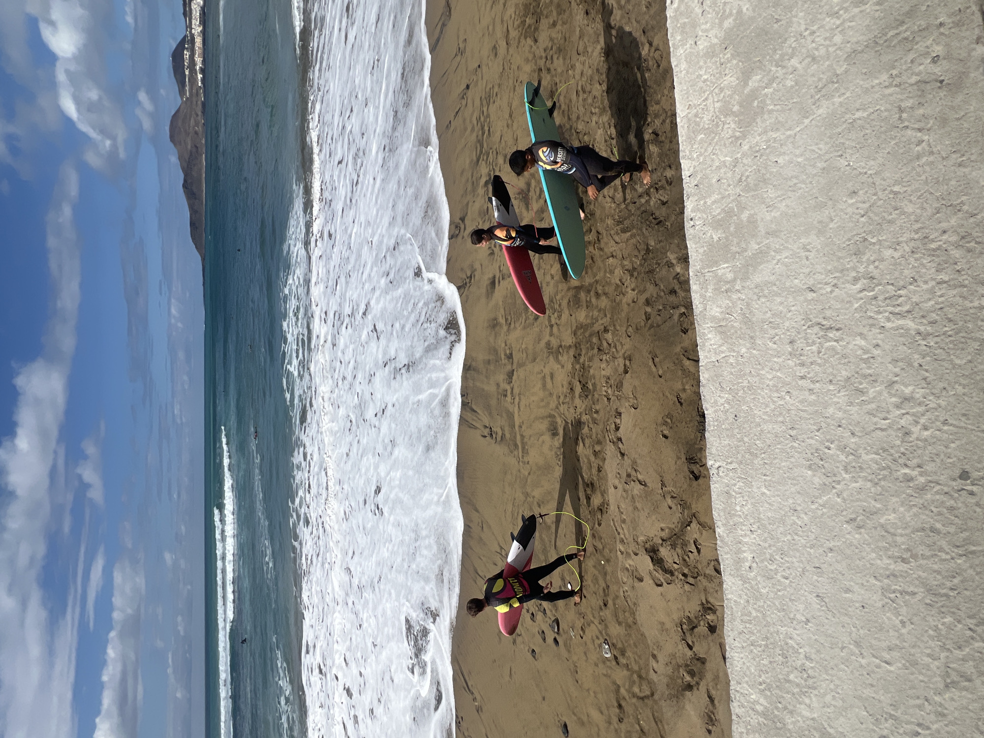 The image displays a sunny daytime scene at a sandy beach in Las Palmas De Gran Canaria, Spain, featuring three individuals with surfboards. The perspective is slightly elevated, looking down towards the ocean and coastline.
In the foreground, a person stands on the wet sand near the water's edge, wearing a dark wetsuit under a bright yellow and pink vest with the text "WAVUTY" visible. They are holding a pink surfboard with a yellow leash attached to their ankle. Further along the beach, two other individuals are present. One wears a dark wetsuit with "UNIVERSITY SURF" printed on their chest and is holding a light blue-green surfboard. Next to them, another person in a dark wetsuit with red accents holds a red surfboard with a black tip. All three appear to be engaged in or preparing for a surfing activity.
The setting includes a wide expanse of light brown sandy beach meeting the clear, blue-green ocean, which features white foam from gentle breaking waves. In the background, large mountains or hills are visible under a bright blue sky dotted with scattered white clouds. To the right of the frame, a grey concrete structure, possibly a walkway or barrier, runs alongside the beach. The bright lighting indicates it is daytime, suggesting conditions are ideal for recreational beach activities.