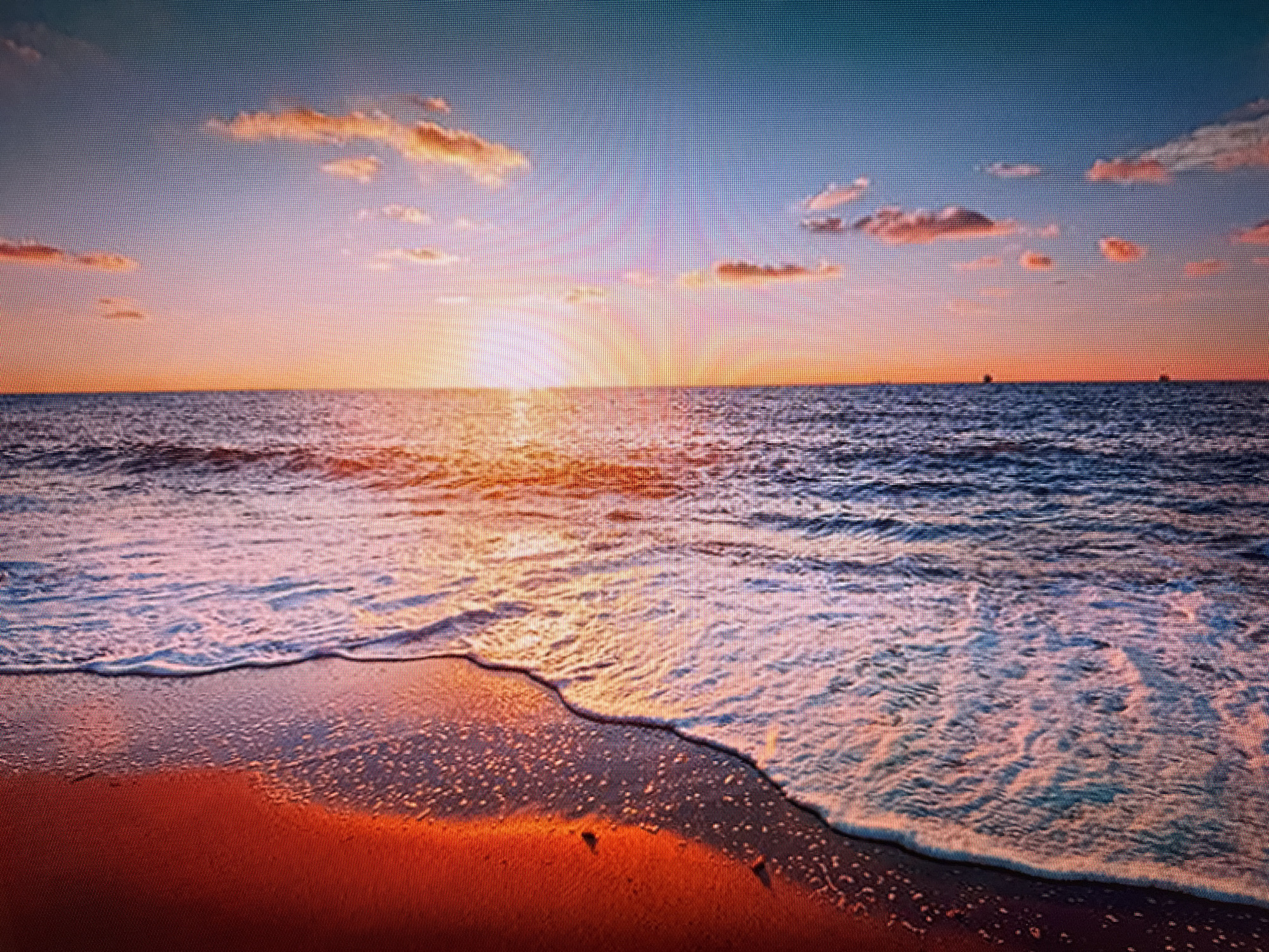 The image depicts a serene sunset over the ocean, with the sun low on the horizon casting a warm, golden glow across the sky and water. The sky transitions from a deep blue at the top to soft hues of pink, orange, and yellow near the horizon, dotted with scattered clouds tinged with the same warm colors. Gentle waves are rolling onto a sandy beach, creating white foam as they reach the shore. The sand itself reflects the sunset colors, appearing reddish-orange in the foreground. Two small, dark shapes, possibly boats or distant landforms, are visible on the horizon. The scene is peaceful and natural, evoking a sense of tranquility. There is no discernible connection to the city of Szczecin, Poland, as the image shows a coastal landscape with no urban elements or recognizable landmarks.