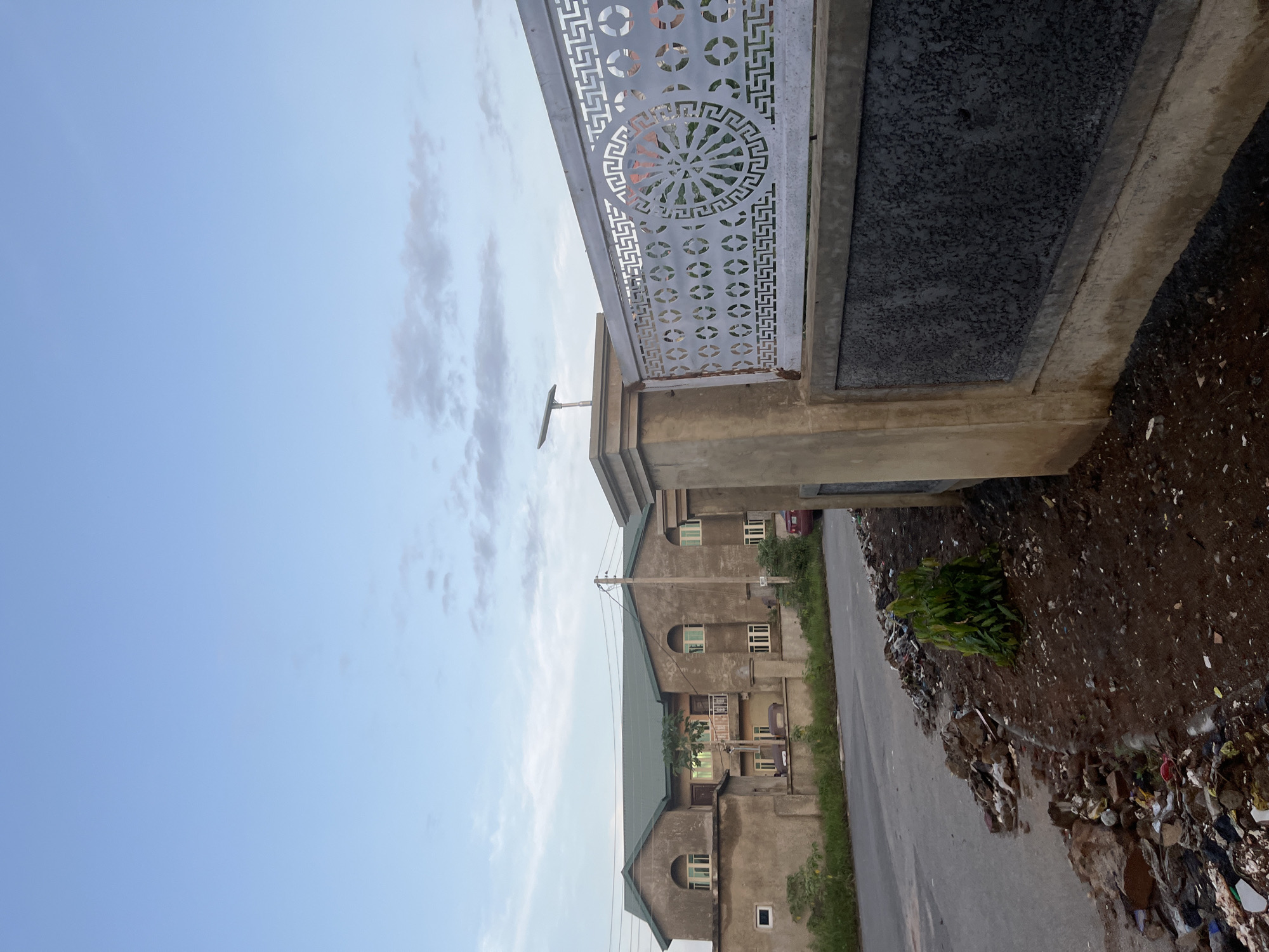 The image depicts a street scene in Abuja, Nigeria. On the right side of the frame, a concrete wall with decorative metal fencing runs along the foreground. The wall has a grey, textured panel inset within a beige concrete structure. The metal fence is white and features intricate geometric patterns and a circular motif. A solar-powered street lamp is visible on top of a concrete pillar behind the fence.
To the left of the wall, a paved road stretches into the background. The roadside is cluttered with debris, including plastic waste, rocks, and other refuse, with a small green plant growing amidst the trash. Further down the road, a large, two-story building with a green roof and beige walls is visible. The building has multiple windows with light-colored frames and arched tops on the upper floor. A red car is parked near the building in the distance. The sky above is a light blue with scattered white clouds.