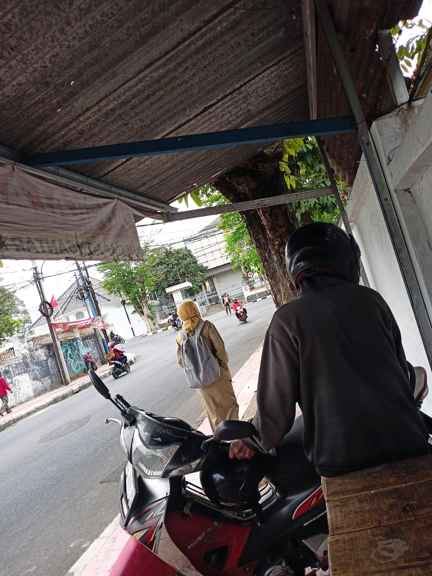 The image depicts a street scene in Magelang, Indonesia, likely during the daytime, under a corrugated metal roof supported by a blue metal beam. In the foreground, a person wearing a dark hooded sweatshirt and a black helmet is seated on a wooden bench, partially obscuring a red and black motorcycle. To the left, another person wearing a yellow hijab and a grey backpack is walking away from the camera, towards a street with several motorcycles and a few pedestrians. The street is paved, with a raised sidewalk on the left side. Buildings with white walls and some architectural details are visible in the background, along with trees and utility poles. The overall atmosphere suggests an urban environment.