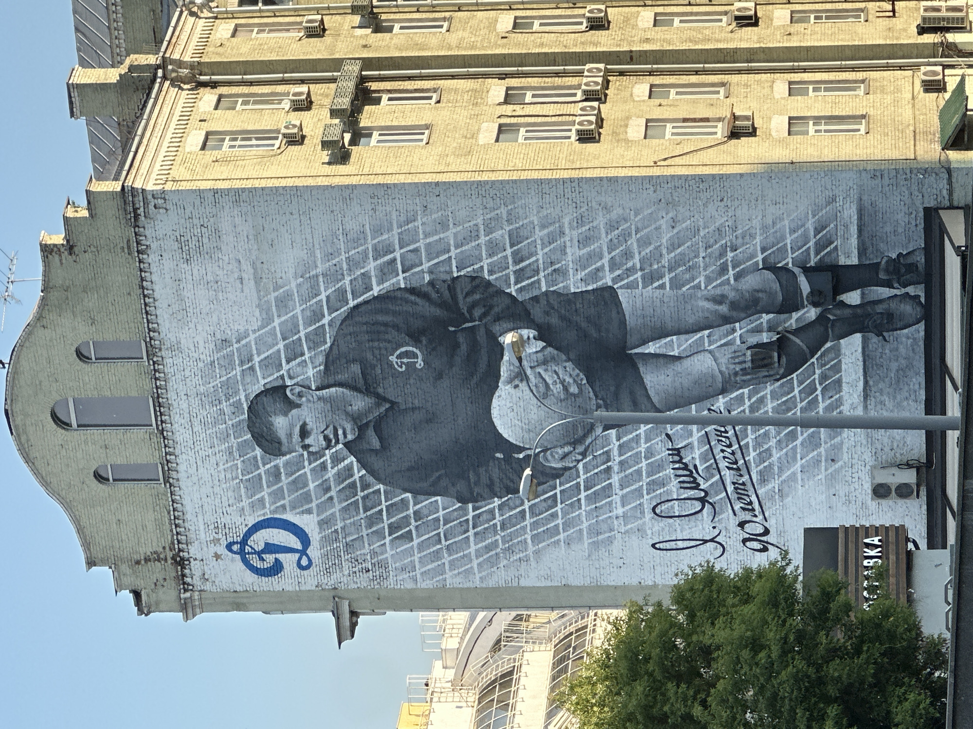 This is a vertical, outdoor photograph of a large mural painted on the side of a building in Moscow, Russia. The mural depicts a black and white portrait of a man in athletic attire, holding a ball. The man is rendered in a realistic style, with a serious expression. The mural is set against a backdrop of a white, diamond-patterned grid. To the left of the portrait, there is a blue logo resembling a stylized "D" with two stars. To the right of the portrait, there is a calligraphic inscription in black. Below the mural, a green tree is visible, and a modern building with a curved facade can be seen in the background.
The building itself appears to be made of yellow brick and has several windows and air conditioning units. The sky is a clear blue, suggesting it is daytime with fair weather. The overall mood of the mural is one of respect and commemoration.
Visible text includes the blue logo and the black calligraphic inscription, which appears to say "Л. И. Яшин 90 лет легенды" which translates to "L. I. Yashin 90 years of legend." Another visible sign on a box-like structure to the right reads "КОФЕЙКА".