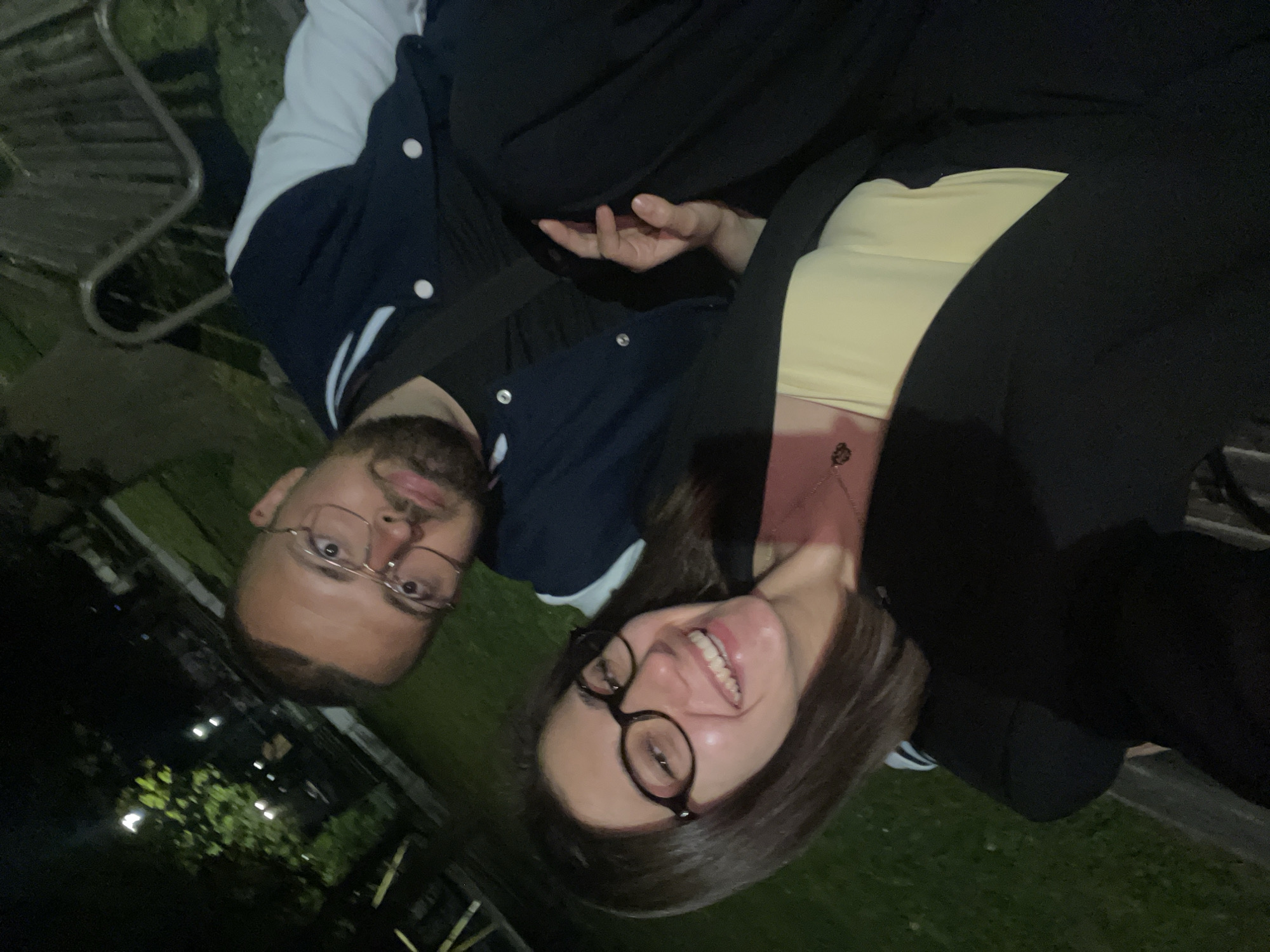The image captures a selfie-style shot of a man and a woman sitting outdoors at night, likely in a park or green area in Sofia, Bulgaria. The original image is rotated 90 degrees clockwise, showing them lying on their sides.
The man, positioned on the left, has short dark hair and a beard, and wears glasses. He is dressed in a dark blue varsity-style jacket with white sleeves and a white collar, over a dark t-shirt. He looks directly at the camera with a slight smile.
The woman, on the right, has dark hair, also wears glasses, and has a broad, cheerful smile, revealing her teeth. She is wearing a black blazer or jacket over a light yellow top, and a delicate necklace is visible around her neck. Her right hand is visible resting near the man's arm.
The background is dark, characteristic of nighttime, with blurry artificial lights scattered among dark foliage, indicating trees or bushes. The ground beneath them appears to be grass. Both individuals exhibit happy and relaxed expressions, suggesting a pleasant interaction or event. There is no visible text in the image.