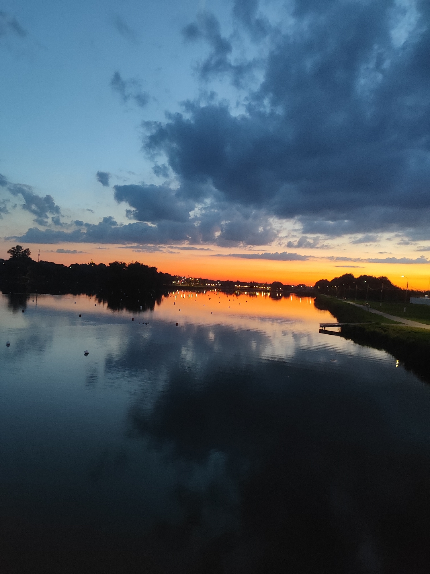 The image captures a sunset over the Bronnitsy Reservoir in Bronnitsy, Russia. The sky is a gradient of blues and grays with dark clouds dominating the upper portion, transitioning to a vibrant orange and yellow band near the horizon. Silhouetted trees and buildings line the far bank of the reservoir. The water's surface reflects the colorful sky and the dark treeline, creating a mirror-like effect. Several buoys are visible in the water, some appearing as white dots. A small wooden pier extends into the water from the right bank, which also features a walking path and streetlights. The overall scene depicts a serene evening at the reservoir.