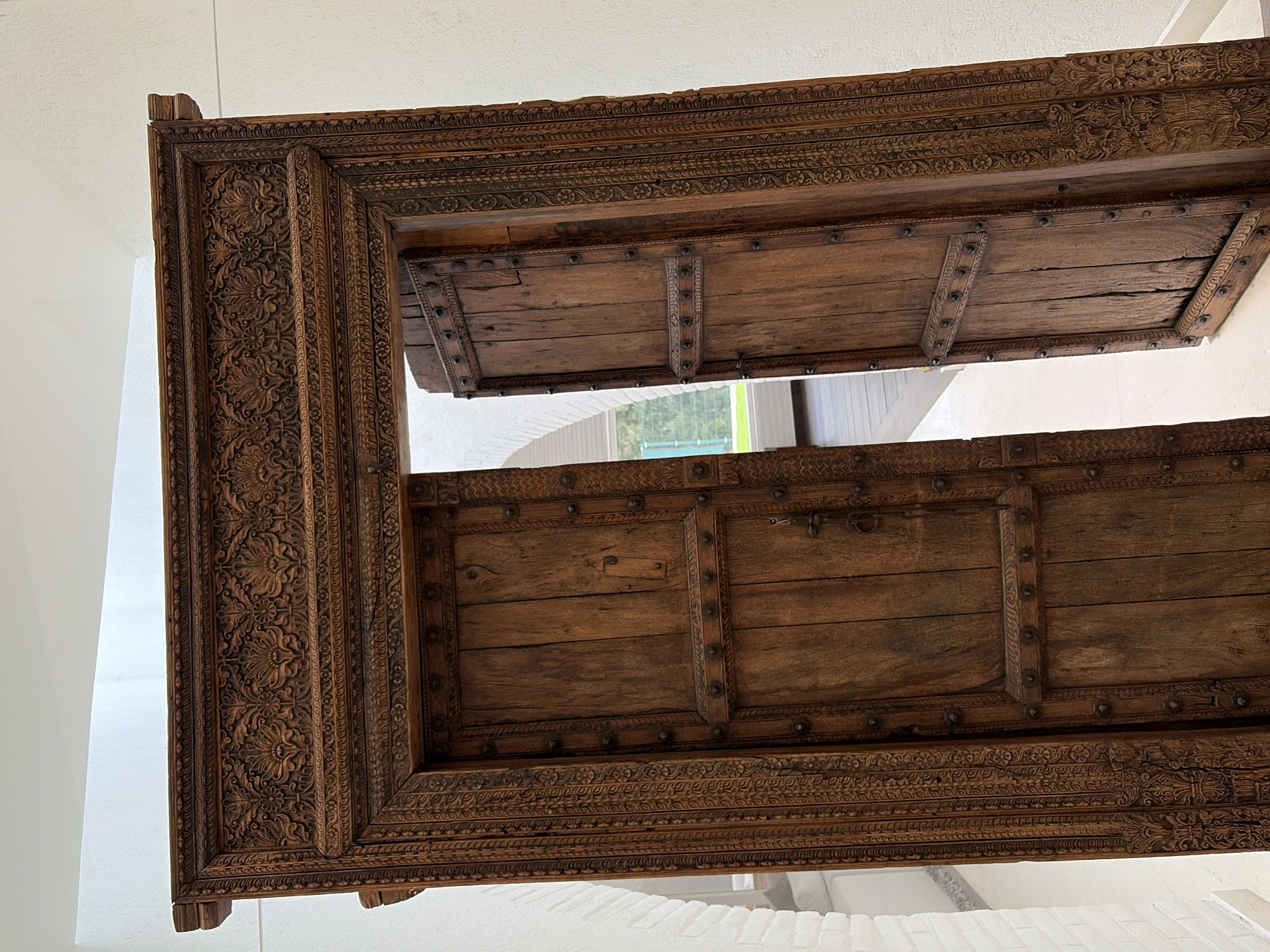 A large, ornate wooden doorway, featuring intricate carvings, is the central subject of the image. The door frame is crafted from dark, rich brown wood, possibly teak, and is extensively carved with complex floral and geometric patterns that run along its vertical and horizontal sections. The carving style suggests a traditional, possibly South Asian, aesthetic.
Two wooden doors, lighter in tone than the frame, are set within this structure. They are each composed of several horizontal planks, secured with vertical metal straps adorned with numerous metal studs or rivets. The wood of the doors shows natural grain, some wear, and minor imperfections, indicating age and character. The top door is partially open, revealing a glimpse of an outdoor setting with green foliage and a modern-looking structure in the distance. The bottom door is also partially ajar, and a metal ring pull or handle is visible on its right panel.
The setting appears to be an indoor or covered space, characterized by plain white walls and ceiling, suggesting a gallery, museum, or well-lit architectural space. The bright lighting indicates it is daytime. No people or explicit activities are depicted, but the open doors imply passage or display. There is no visible text in the image. The object is situated in what appears to be San Bernardino Tlaxcalancingo, Mexico.