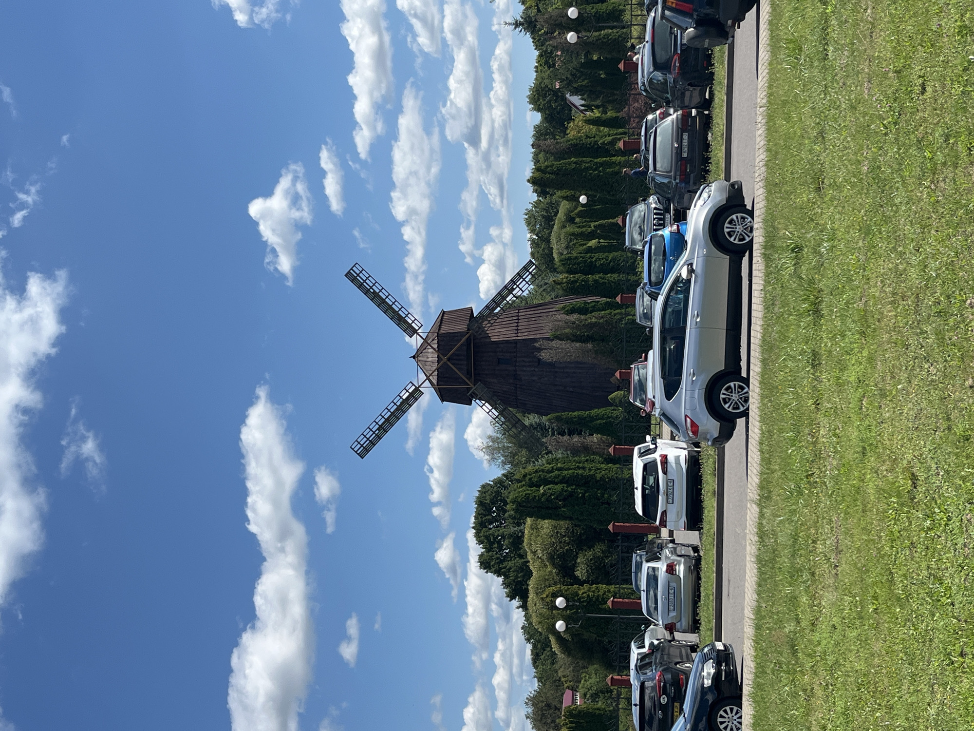 An outdoor daytime, medium-wide shot captures a parking lot with various cars in front of a traditional wooden windmill. The windmill, constructed of dark wood, has four prominent, slatted blades angled against a clear blue sky dotted with white clouds. The surrounding area is lush with green trees and bushes. A paved road separates the parking lot from a grassy foreground. Several cars are parked in the lot, including a silver SUV in the foreground, and a line of cars behind it. The sky is a vibrant blue with fluffy white clouds. The scene appears to be located in Буйніцкі сельскі Савет, Belarus.