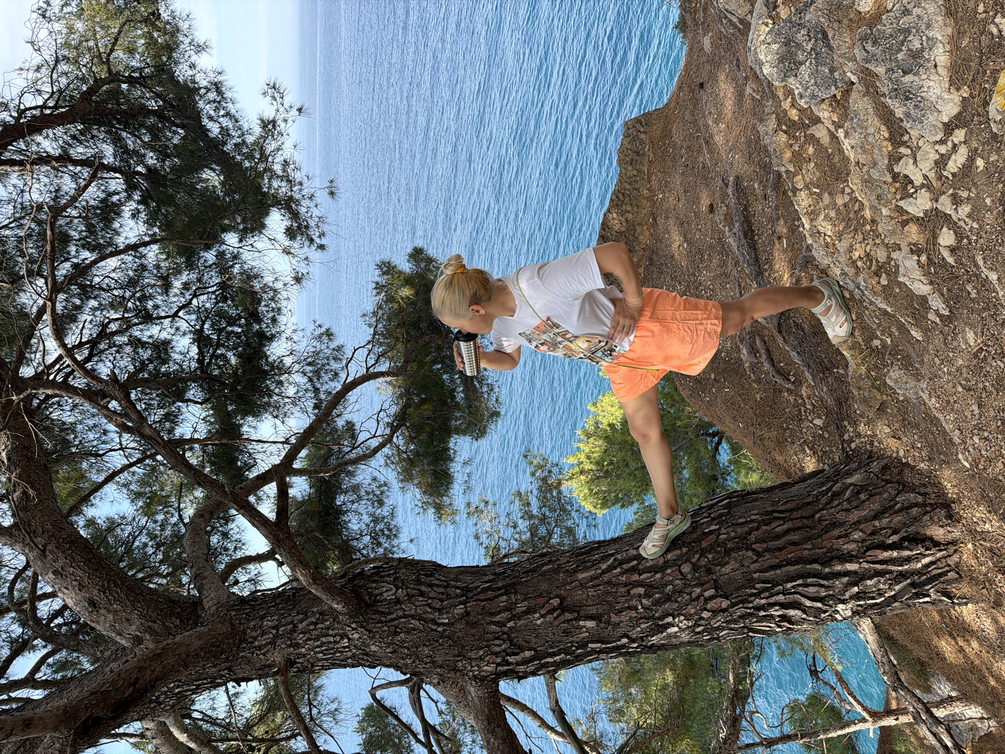 A woman stands on a tree trunk in Petrovac na Moru, Montenegro, drinking from a metal flask. The tree trunk spans out over a cliff with the sea visible below. The woman has blonde hair in a bun and wears a white t-shirt and orange shorts, with sandals on her feet. The sea is blue and calm, and parts of the rocky cliff are visible. The tree is bare with many branches. The camera is tilted.