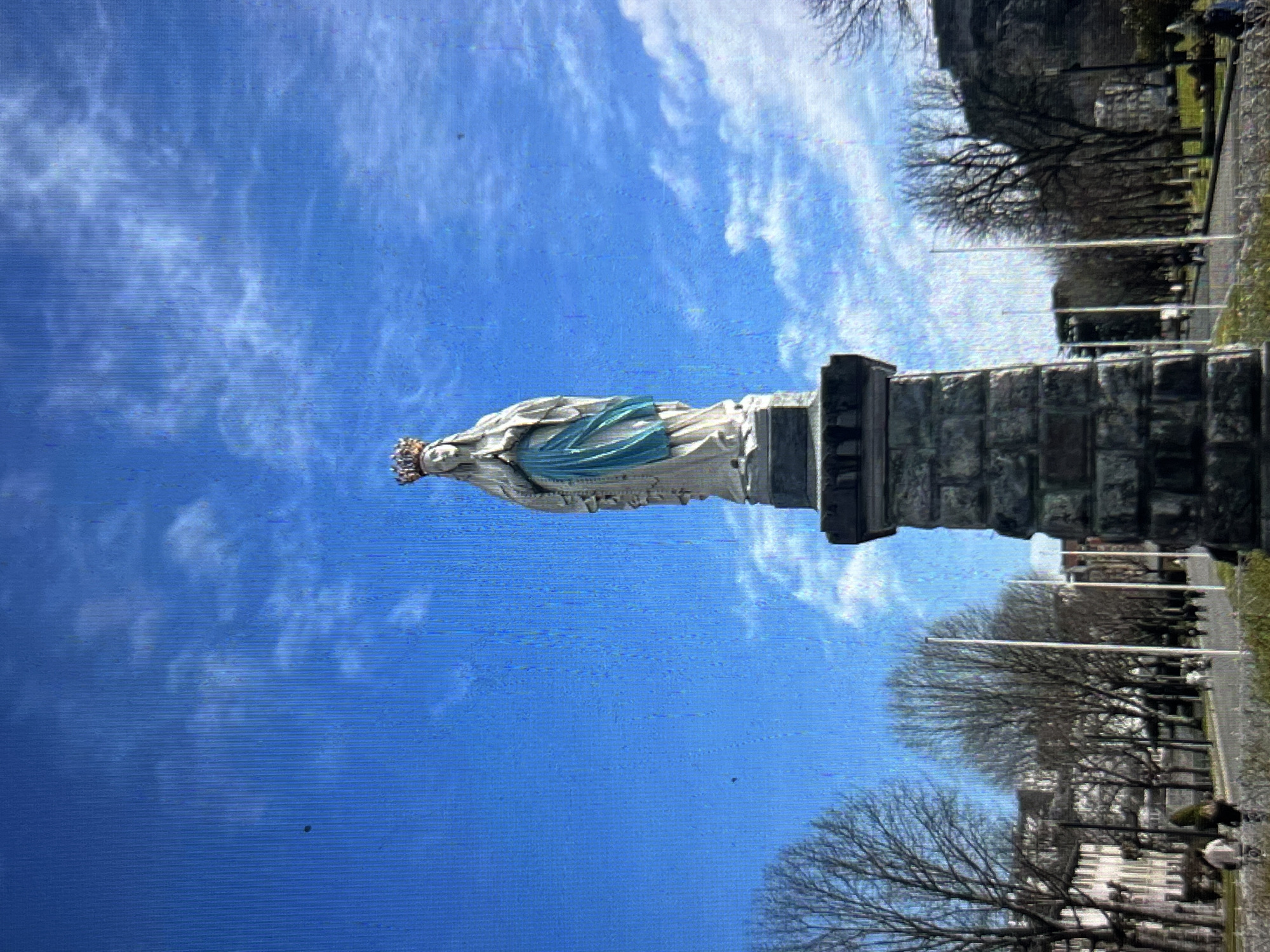 The image shows a statue of the Virgin Mary standing on a stone pedestal. The statue is white with a blue sash. A crown is on her head. The sky behind the statue is blue with white clouds. To the right of the statue are trees and a street. This is the Virgin Mary statue in the city of Tinaquillo, Venezuela.