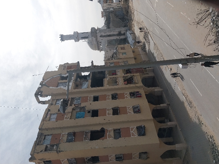 The image displays a desolate urban street scene exhibiting widespread destruction, likely the aftermath of a conflict or major disaster. The photograph itself is significantly tilted, appearing as if the buildings are leaning and the street runs steeply uphill from left to right.
In the foreground and mid-ground, a multi-story building, possibly residential or commercial, dominates the left side of the frame. It is constructed from a light-colored material, possibly beige or light brown, with darker contrasting accents. This building is heavily damaged; large sections of its facade are missing, exposing the building's interior. Numerous windows are shattered or entirely absent, leaving dark, empty voids. Some window openings are covered with blue tarps or cloths. Several balconies are visible, many also showing signs of damage or containing debris. The upper floors appear particularly devastated, with visible structural collapse.
A wide, paved road runs through the center and right side of the image, extending into the background. While the road surface appears somewhat cleared, its edges are lined with rubble, dirt, and construction debris. On the far right, a substantial pile of twisted metal rebar and other structural wreckage is visible.
A thick, dark utility pole or structural beam crosses the image horizontally in the mid-ground, spanning over the road. Wires are strung from it, and a white rectangular box is attached to its underside.
In the background, further down the street, stands a large building that strongly resembles a mosque, identified by its distinctive white minaret and dome. This structure also exhibits significant damage, with parts of the minaret and other sections appearing to be broken or collapsed. Other damaged buildings are partially visible behind and around the mosque.
Several small human figures are scattered along the road in the mid-ground and background. They appear to be walking or standing, navigating the debris-strewn environment, suggesting people are moving through or surveying the damaged area.
The sky above is uniformly bright and overcast, indicating daytime conditions without direct sunlight. The overall atmosphere conveyed by the image is one of devastation, desolation, and the grim aftermath of a destructive event. There is no discernible text visible anywhere in the image.