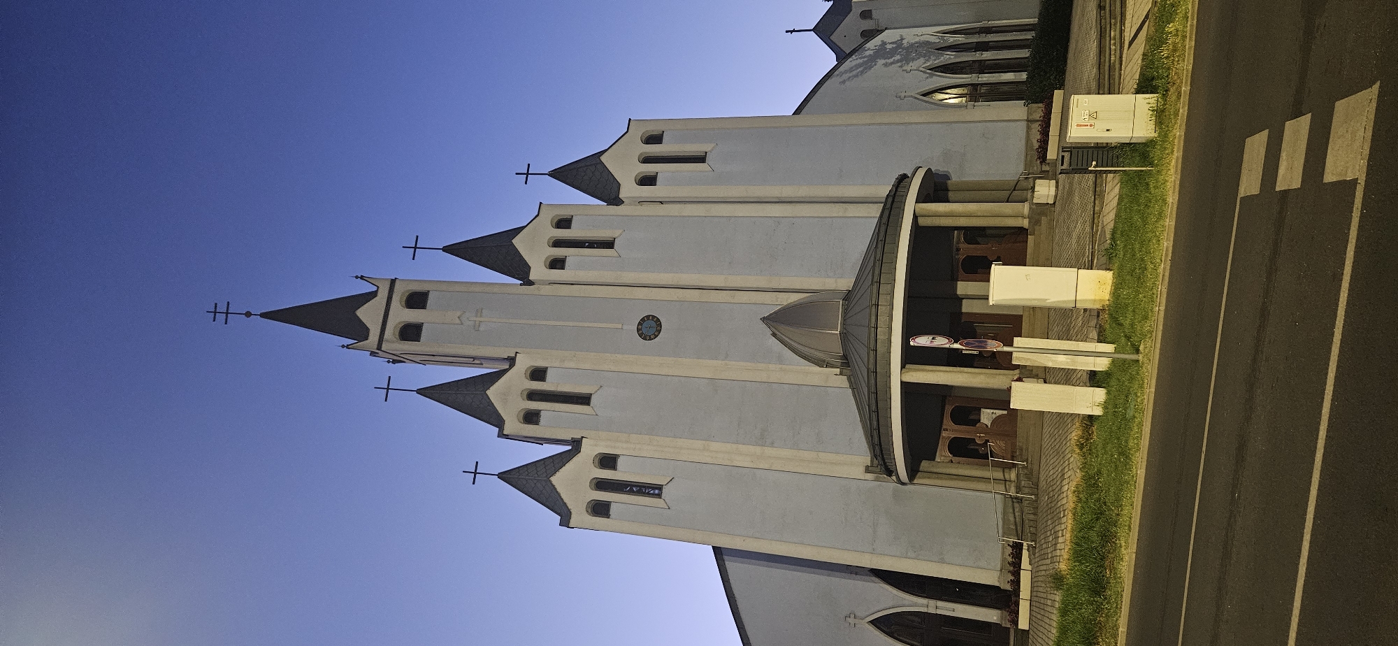 The image shows the Saint Elisabeth of Hungary Church in Hévíz, Hungary, at dusk. The church is a modern Roman Catholic church with a prominent, light-colored facade, topped with several spires, each adorned with a cross. The central tower is the tallest, featuring a clock face. The entrance is accessed via a covered walkway with a rounded roof supported by columns. The building is situated next to a road, with a grassy area and some utility boxes in the foreground. The sky is a deep blue, indicating twilight. There are no people visible in the image.