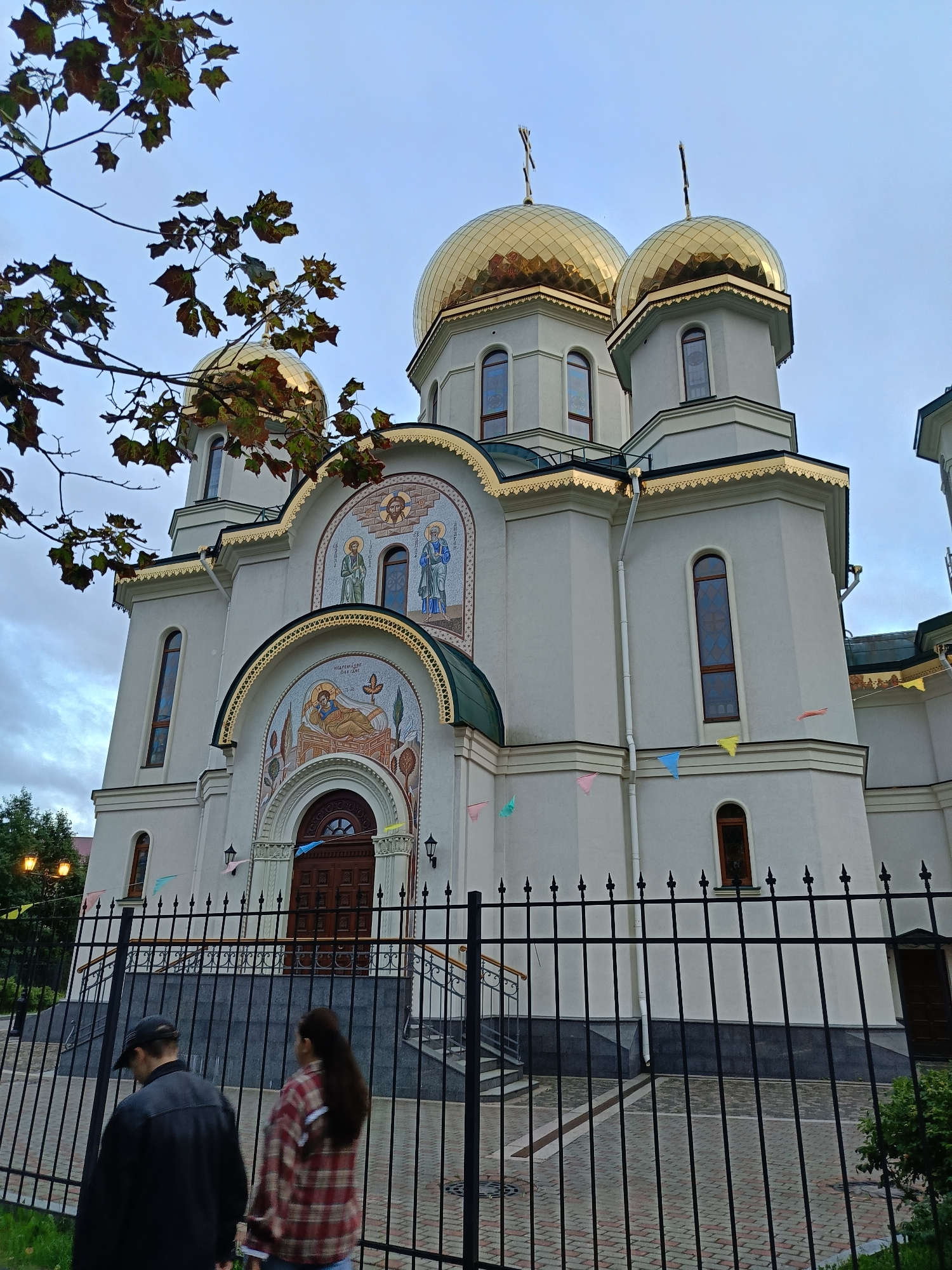 The image shows an exterior view of a white Orthodox church with golden domes against a cloudy sky. The church features intricate mosaic art above its arched wooden entrance. A black metal fence stands in the foreground, with a paved walkway and some greenery behind it. Two individuals, a person in a black jacket and baseball cap, and another person in a red and white plaid shirt, are walking away from the camera, towards the church entrance, on the paved walkway behind the fence. Small, colorful triangular flags are strung across the front of the church. In the upper left corner, tree branches with some brown leaves are visible, suggesting the scene takes place during autumn.