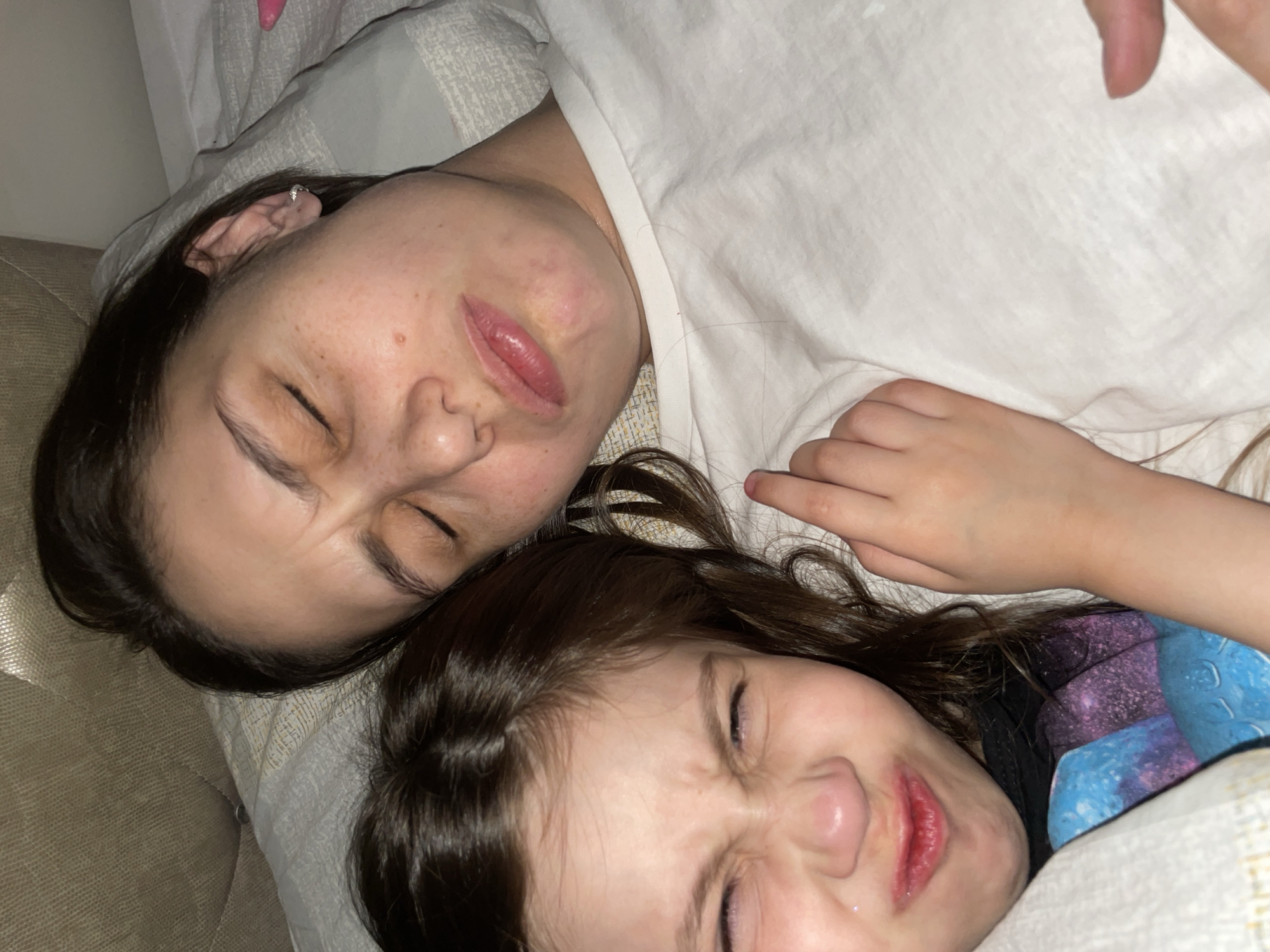 An indoor close-up photograph depicts two female individuals, an adult and a young child, positioned side-by-side on a bed. The specific location within Izhevsk, Russia, cannot be determined from the visual content of the image.
The adult female, with dark hair and fair skin exhibiting freckles, occupies the right side of the frame. She is lying on a light-colored pillow, wearing a plain white t-shirt. Her eyes are closed, her eyebrows are furrowed, and her lips are pursed in a pronounced grimace. A small silver stud earring is visible in her right ear.
The young female child, with dark brown hair and fair skin, is positioned to the left of the adult. She is lying closely alongside the adult, partially covered by a white patterned sheet or blanket. Her eyes are tightly squeezed shut, her nose is scrunched, and her lips are pursed, mirroring the grimacing expression of the adult. A dark-colored garment with blue and purple patterns is visible beneath the sheet. The child's left hand is visible, with a finger touching the adult's upper body.
In the background, a textured, light-colored headboard is partially visible above the individuals' heads. The overall lighting is consistent with an indoor setting.
