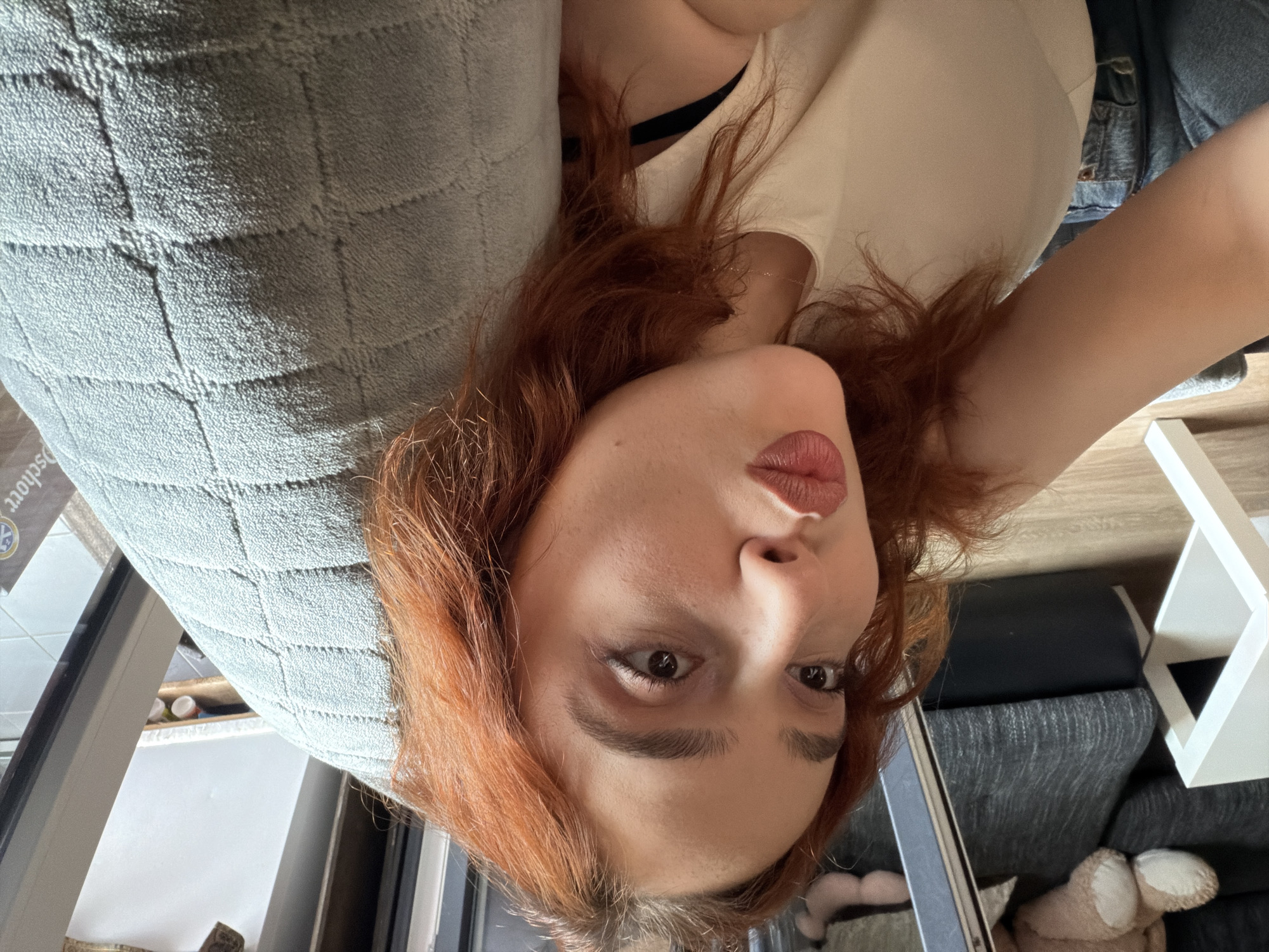 A young woman with reddish-brown hair is depicted in a close-up selfie, oriented as if lying down and looking up at the camera. She has light skin, defined eyebrows, and dark eyes, with her lips slightly pursed. She is wearing a light-colored top, possibly beige or off-white, with a dark bra strap visible near her shoulder. Her head rests on a grey, plush pillow or cushion with a quilted, rectangular pattern.
In the background, on the right side of the image, a dark grey sofa or armchair is visible, along with a white, geometric decorative object and a light brown teddy bear or plush toy. On the left side, there's a glimpse of a window frame and an indoor setting with what appears to be shelves or furniture in the distance. The lighting suggests an indoor environment, possibly during the daytime, with soft, ambient light. The overall setting appears to be a living room or bedroom. The woman's expression is neutral, bordering on playful. Part of the word "schorr" is faintly visible on the grey pillow. The location context provided is Strasbourg, France.