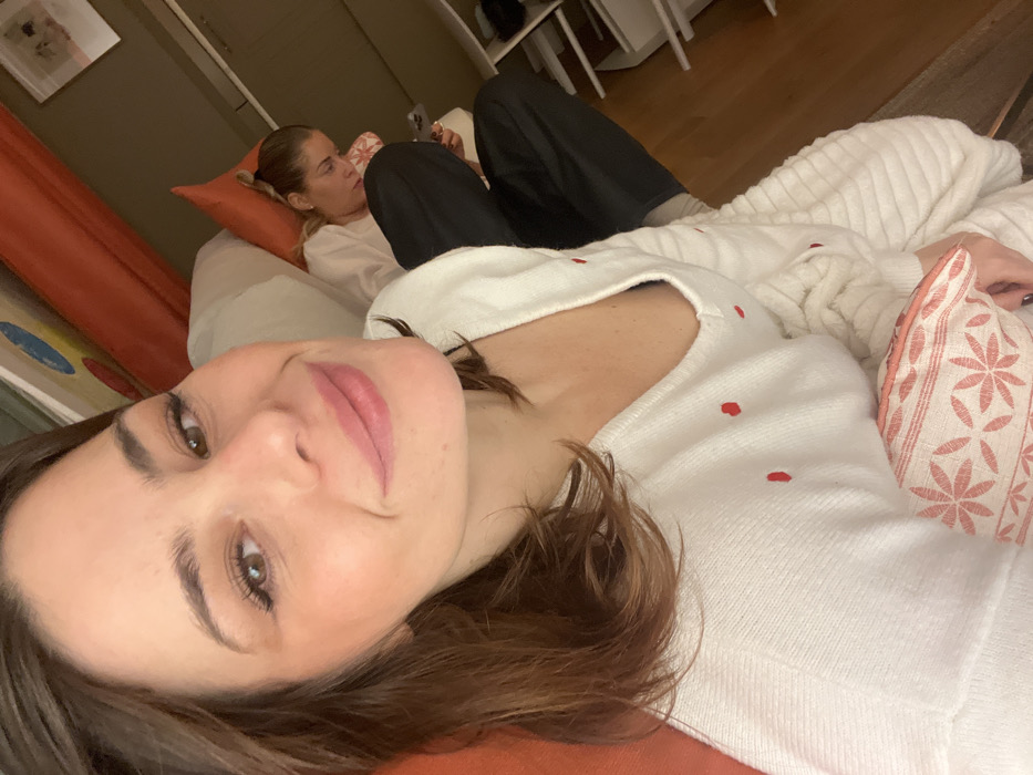 Two women are lounging indoors. The foreground woman is lying on her side, wearing a white sweater with red dots. She has light brown hair and is facing the camera. Behind her, the other woman is reclined on a pillow, wearing a white shirt and black pants. She appears to be looking at something in her hands. The background includes a wooden floor and a glimpse of furniture. The scene has a relaxed and casual atmosphere.