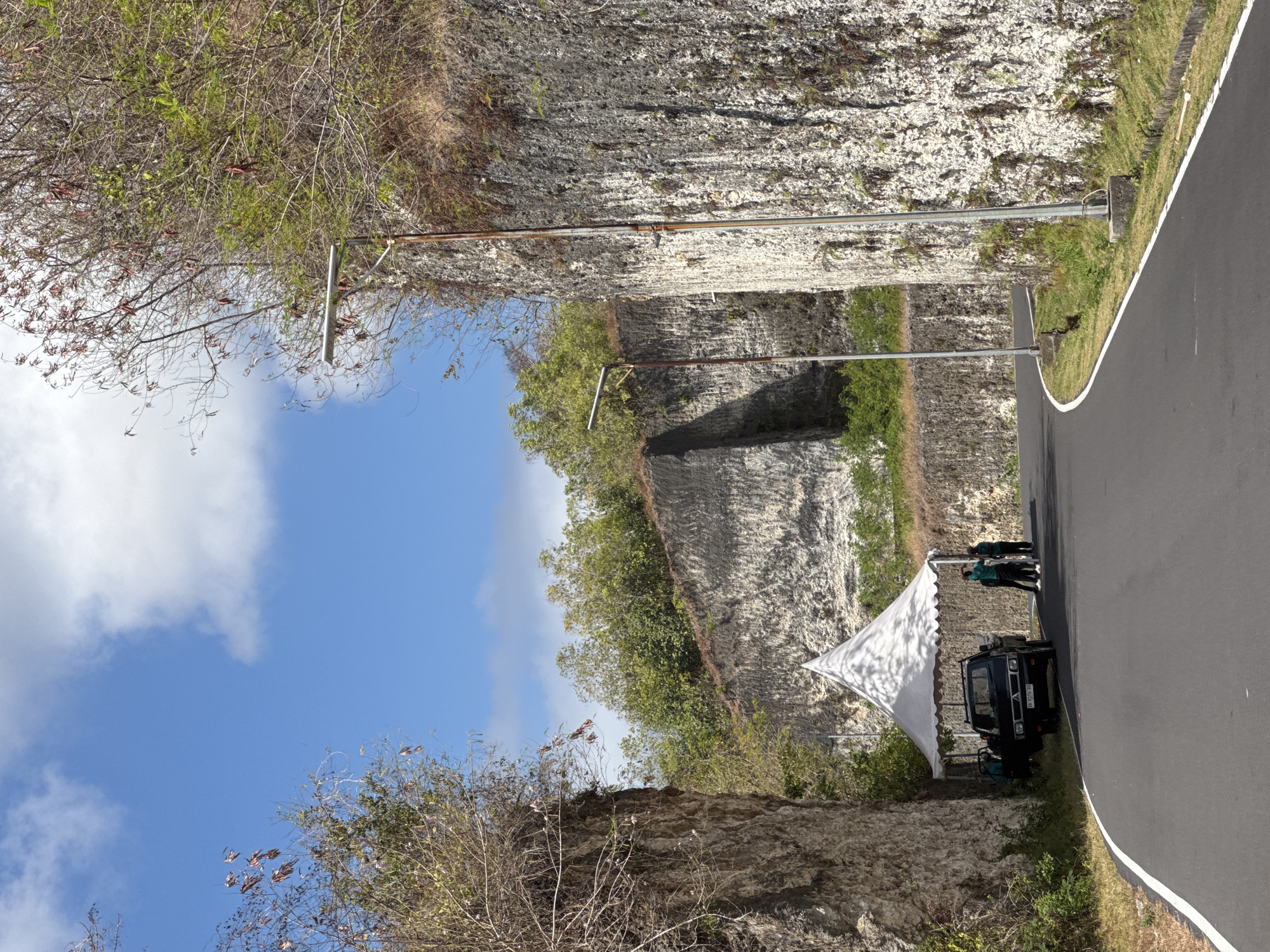A paved road curves through a canyon-like landscape, bordered by steep, rocky cliffs. The cliffs are weathered and have sparse vegetation growing from their surfaces. Two tall streetlights with solar panels stand along the right side of the road. In the distance, on the left side of the road, a white canopy tent is set up next to a dark-colored pickup truck. Three individuals, two wearing green shirts and black pants, and one in a dark top and pants, are near the tent and truck. The sky is blue with scattered white clouds. The scene appears to be located in Kuta Selatan, Indonesia, a region known for its dramatic limestone cliffs and coastal scenery.