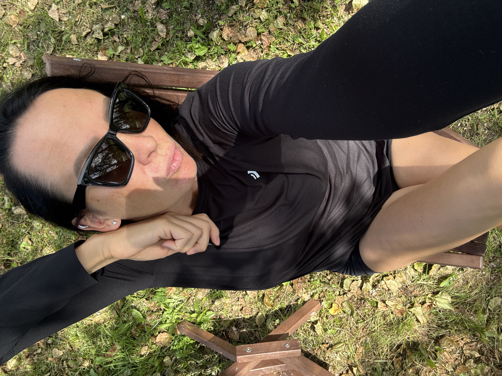 A woman with dark hair and sunglasses is lying on a wooden bench in a grassy area, likely a park or backyard in Chaikovsky, Russia. She is wearing a black long-sleeved shirt and black shorts. Her left arm is resting behind her head, and her right arm is extended, holding a phone to take a selfie. The ground is covered with grass and fallen leaves, indicating it might be autumn. The sunlight casts shadows on her face and body.