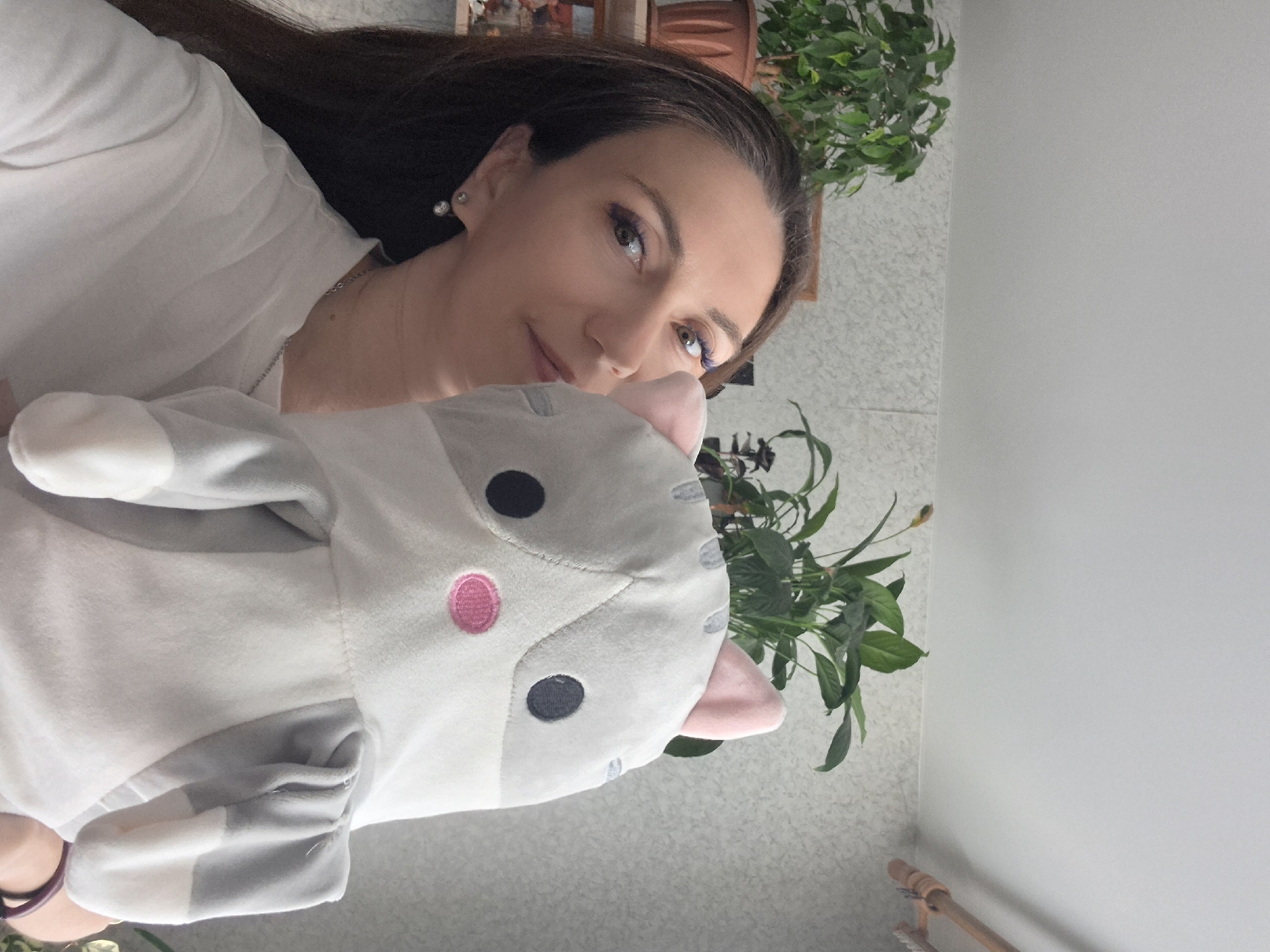 A woman, likely in her late 20s to early 30s, is partially visible in the foreground of the image, holding a stuffed cat toy in front of her face. She has long, dark brown hair, and is wearing a white t-shirt and a silver chain necklace. She has on eye makeup with purple eyeliner. The stuffed cat is predominantly white and grey, with pink ears and a pink nose. Behind the woman, several potted plants are visible on shelves. The background appears to be an indoor setting with a textured, light-colored wall and a white ceiling. A wooden clothes hanger is visible in the upper right corner. The scene is illuminated by natural light, suggesting it was taken during the daytime. The specific location within the city of Chaikovsky, Russia cannot be determined from the visual information provided in the image.